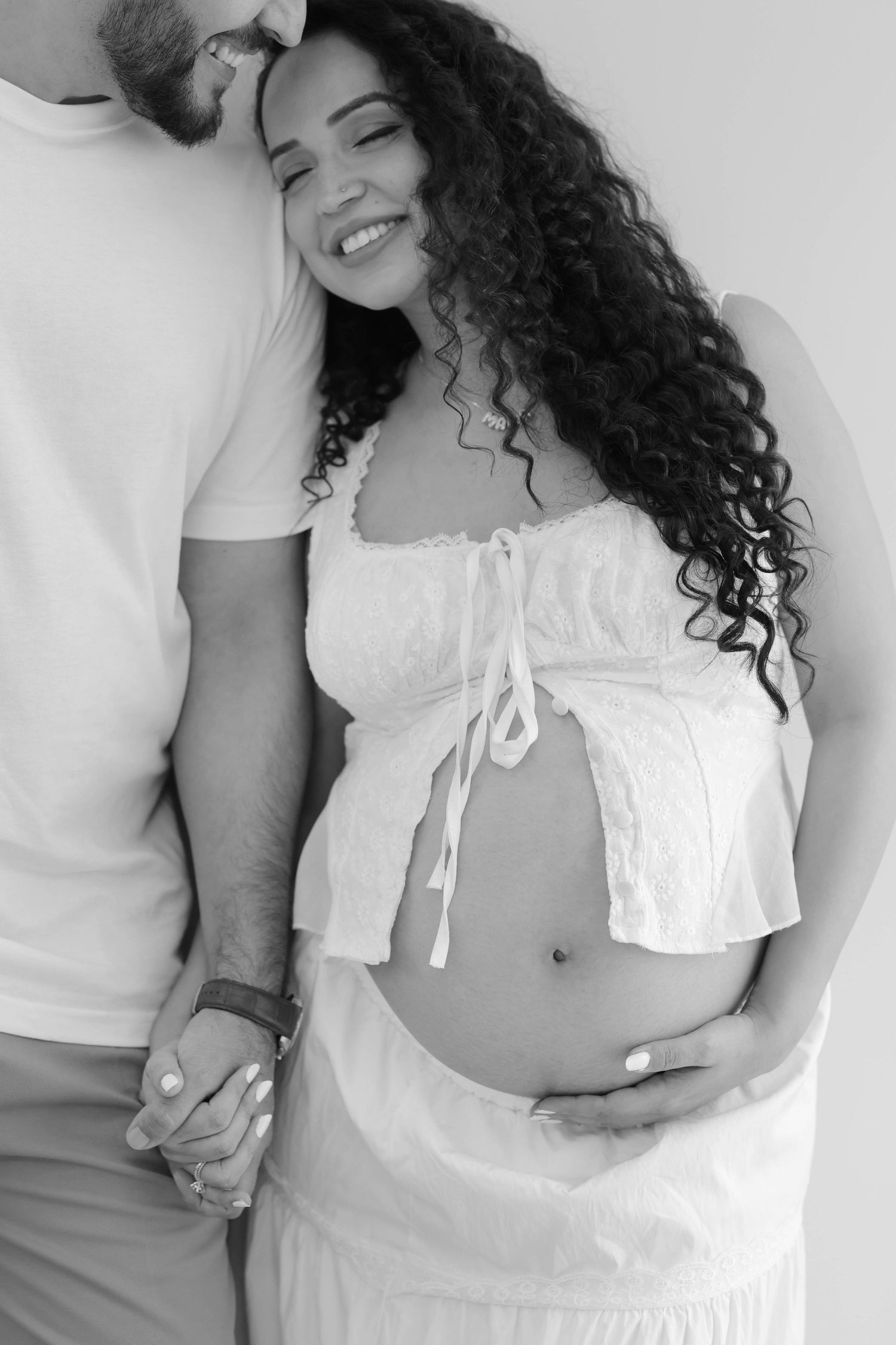 Black and white maternity photo of a smiling pregnant woman in white crop top held by her partner.