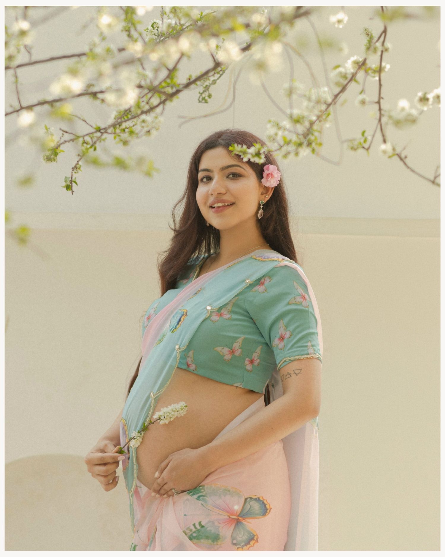 Pregnant woman in pink saree and teal blouse with floral hair accessories poses under blooming white flowers.