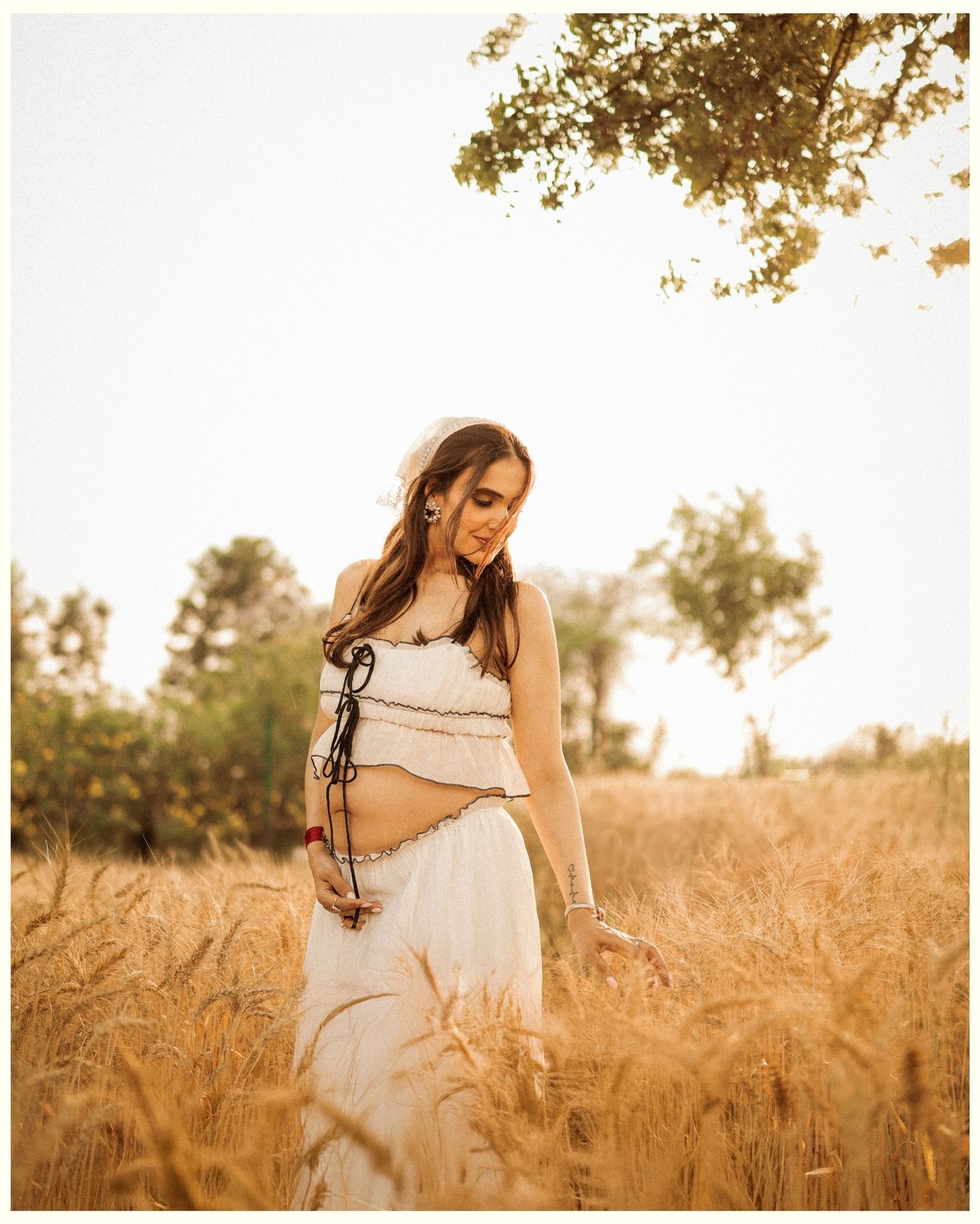 Pregnant woman in white crop top and skirt standing in golden wheat field with warm sunlight and trees in background.
