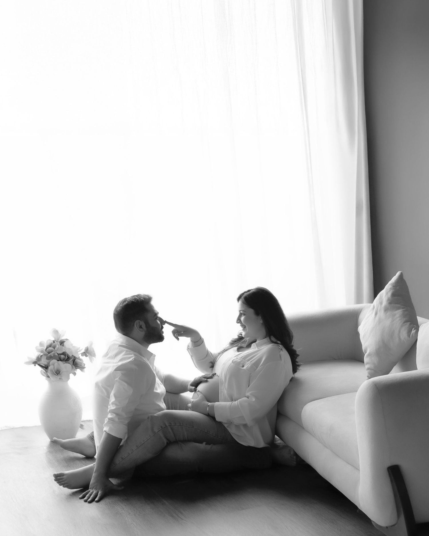 Black and white photo of a couple sitting on the floor by a sofa, sharing a tender moment near a window.