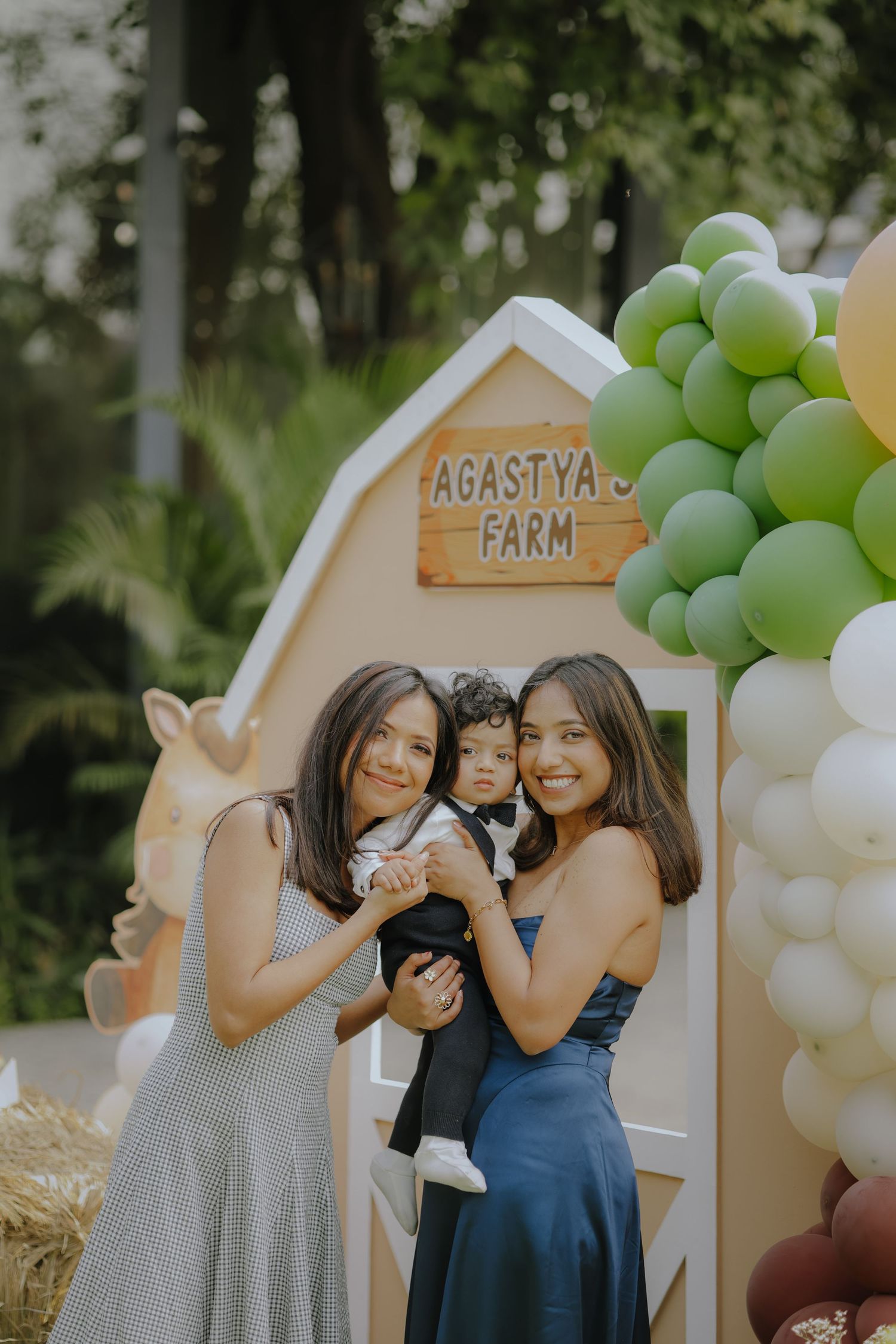 Two women holding a baby boy at Agastya Farm birthday party with green and white balloon decorations.