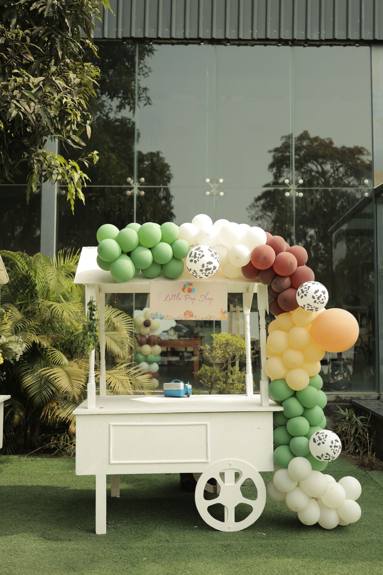 White candy cart decorated with green, white, burgundy, and yellow balloon garland arch outdoors at a party event.