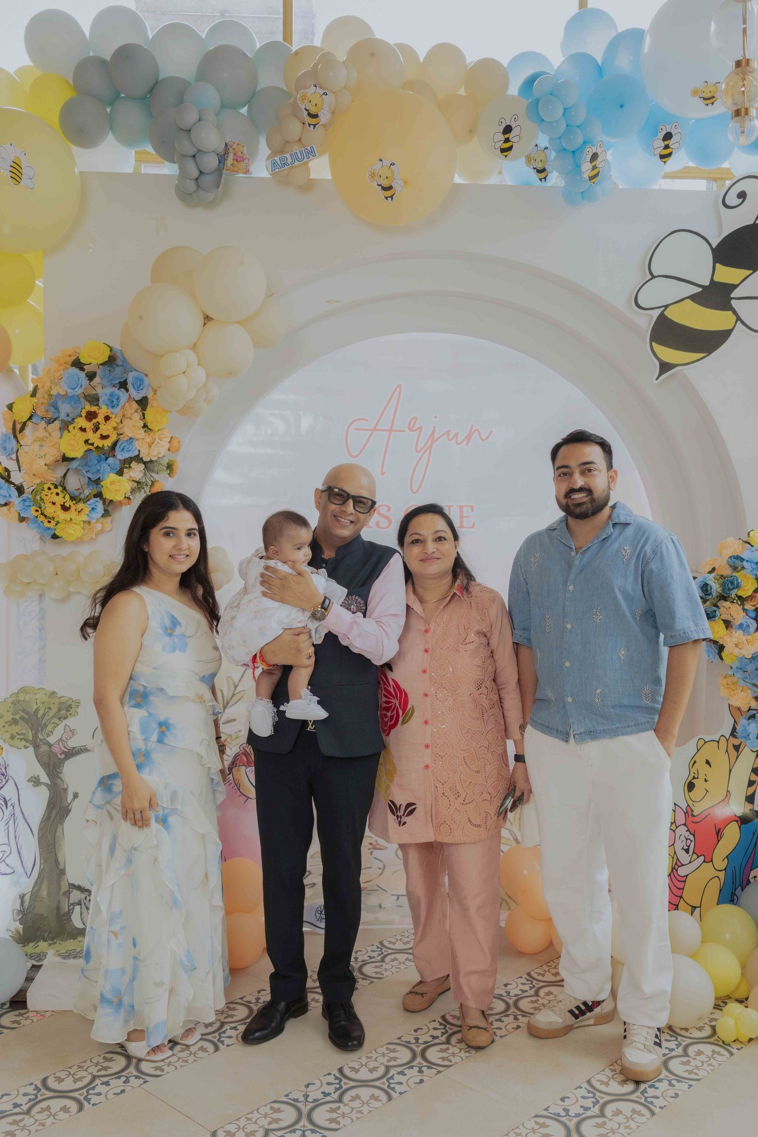 Family posing together at a baby's first birthday party with bee-themed balloon decorations and Winnie the Pooh backdrop.