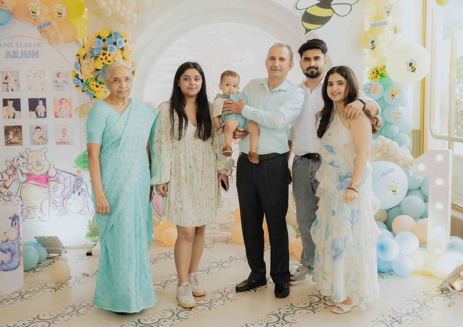 Family posing together at a baby\u2019s first birthday party with balloon decorations and a bee theme backdrop.