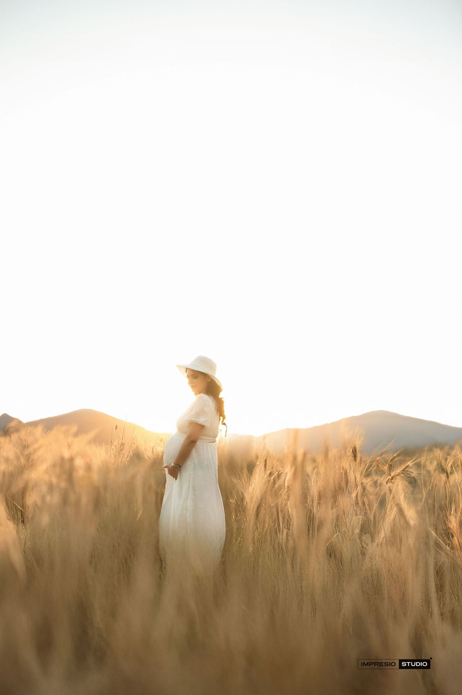 Woman in white dress and hat standing in golden wheat field at sunset with mountains in background.