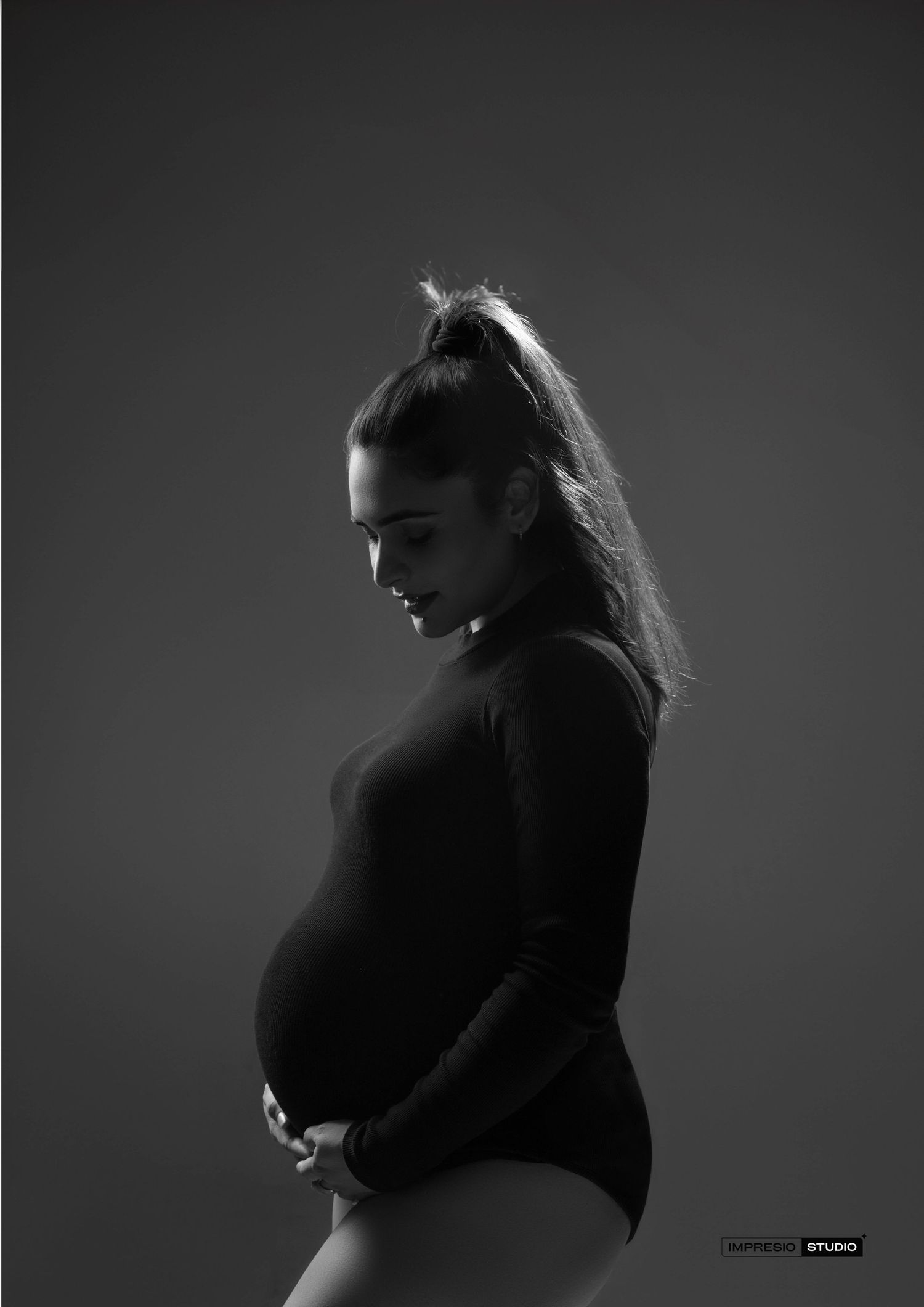 Black and white maternity photo of a pregnant woman in a turtleneck bodysuit cradling her baby bump in a studio.