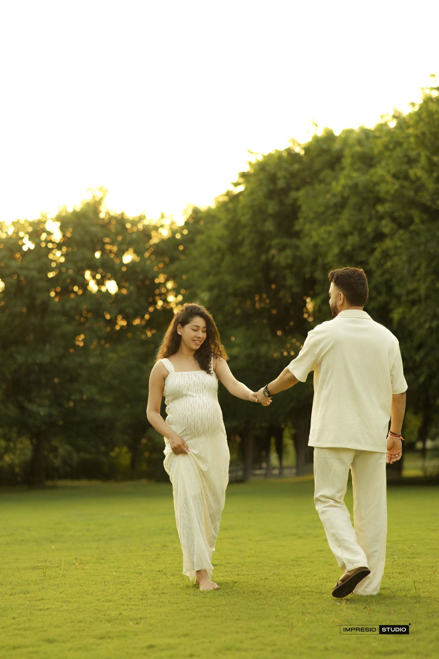 Pregnant woman in white dress holds hands with man in cream outfit during golden hour maternity photoshoot in lush green park.