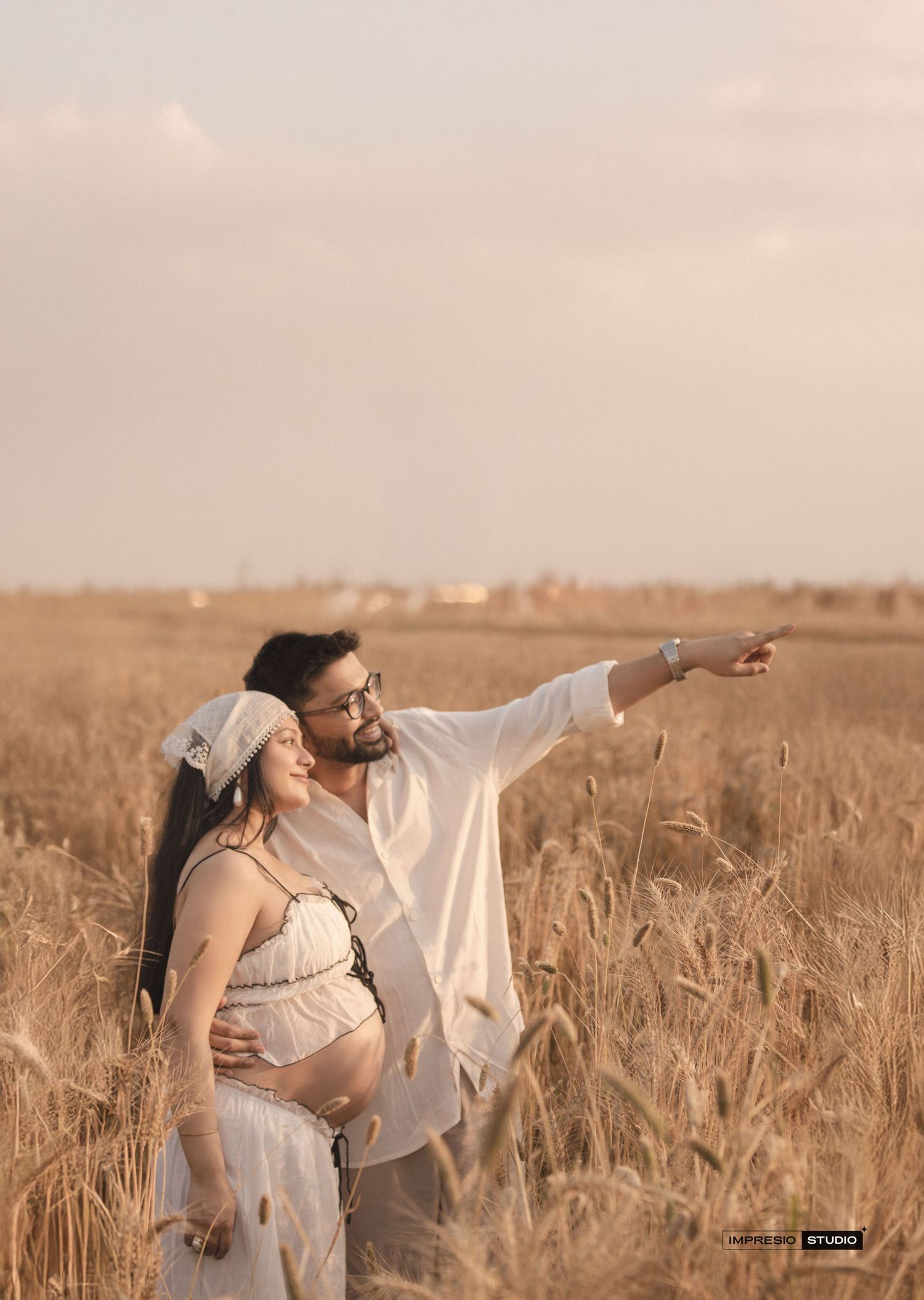 A couple poses in a wheat field, the man pointing to the horizon while embracing his pregnant partner.