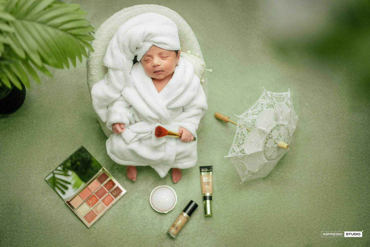 Newborn baby in white robe posed with makeup products and umbrella on green background.