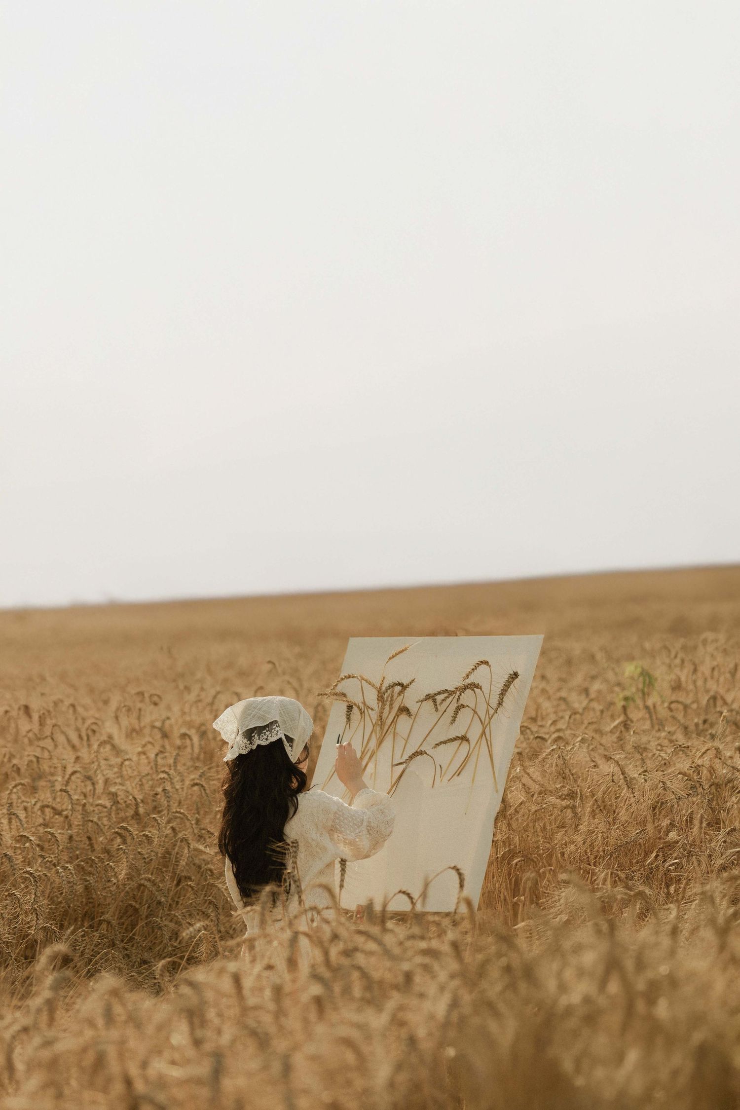 A woman with dark hair paints wheat stalks on a canvas easel in a golden wheat field under an overcast sky.