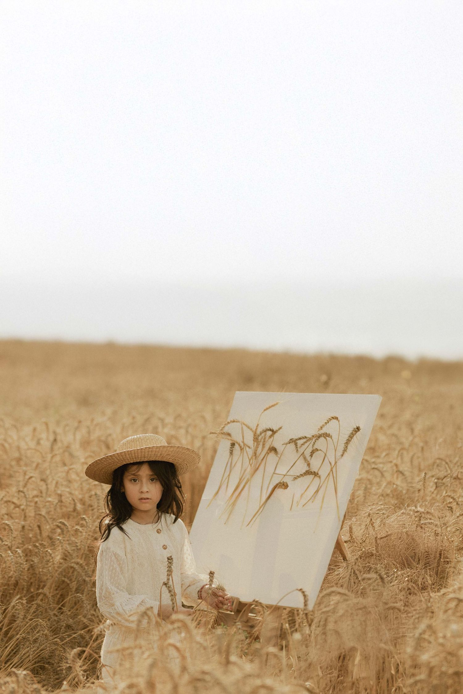 Young girl in straw hat painting wheat stalks on canvas easel in golden wheat field.