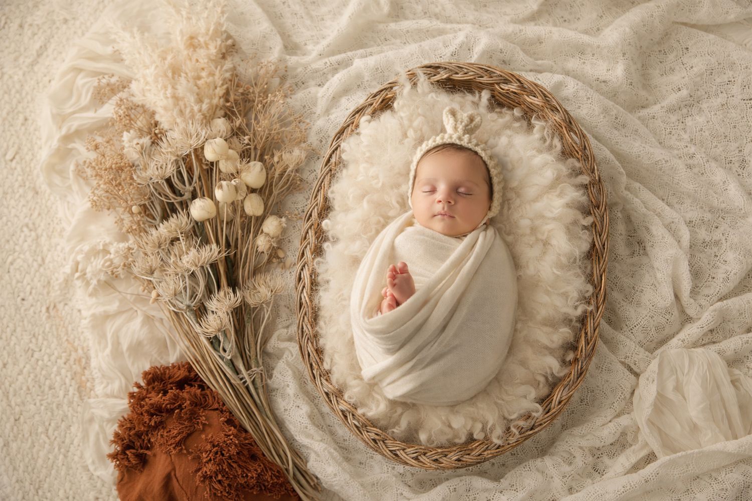 Newborn baby wrapped in white swaddle sleeping in wicker basket with boho floral decor on cream backdrop.