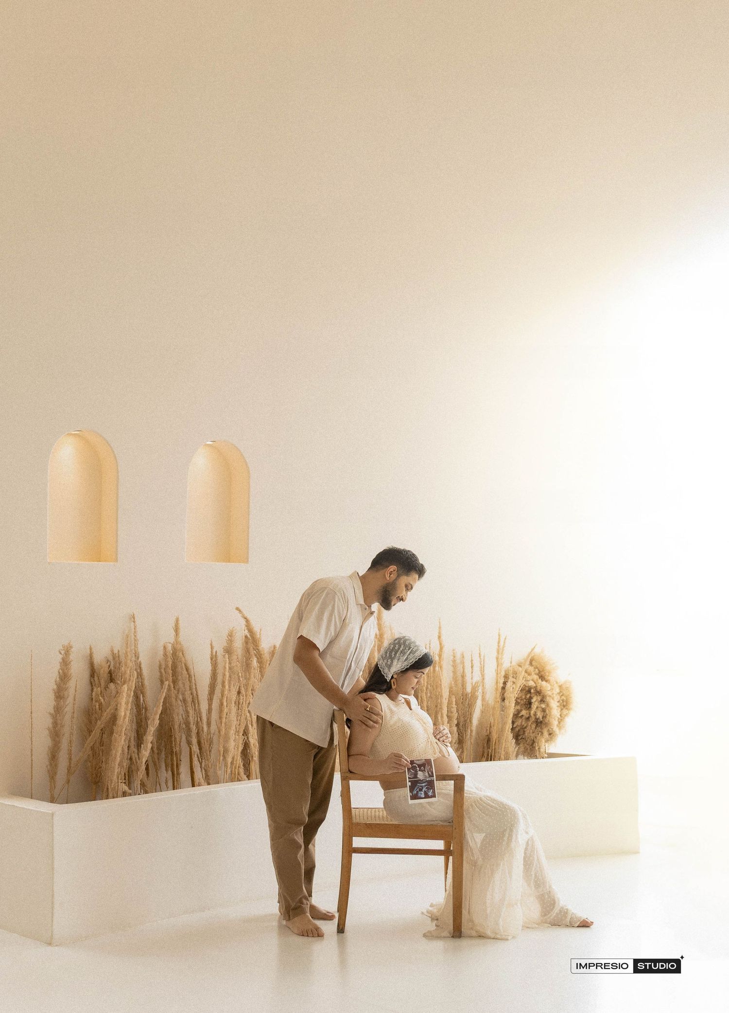 Couple sharing a tender maternity moment in a minimalist beige studio with dried pampas grass decor.