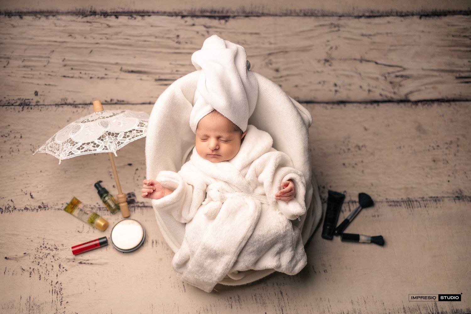 Newborn baby in white robe and towel wrap posed in bowl with miniature spa accessories for photography.