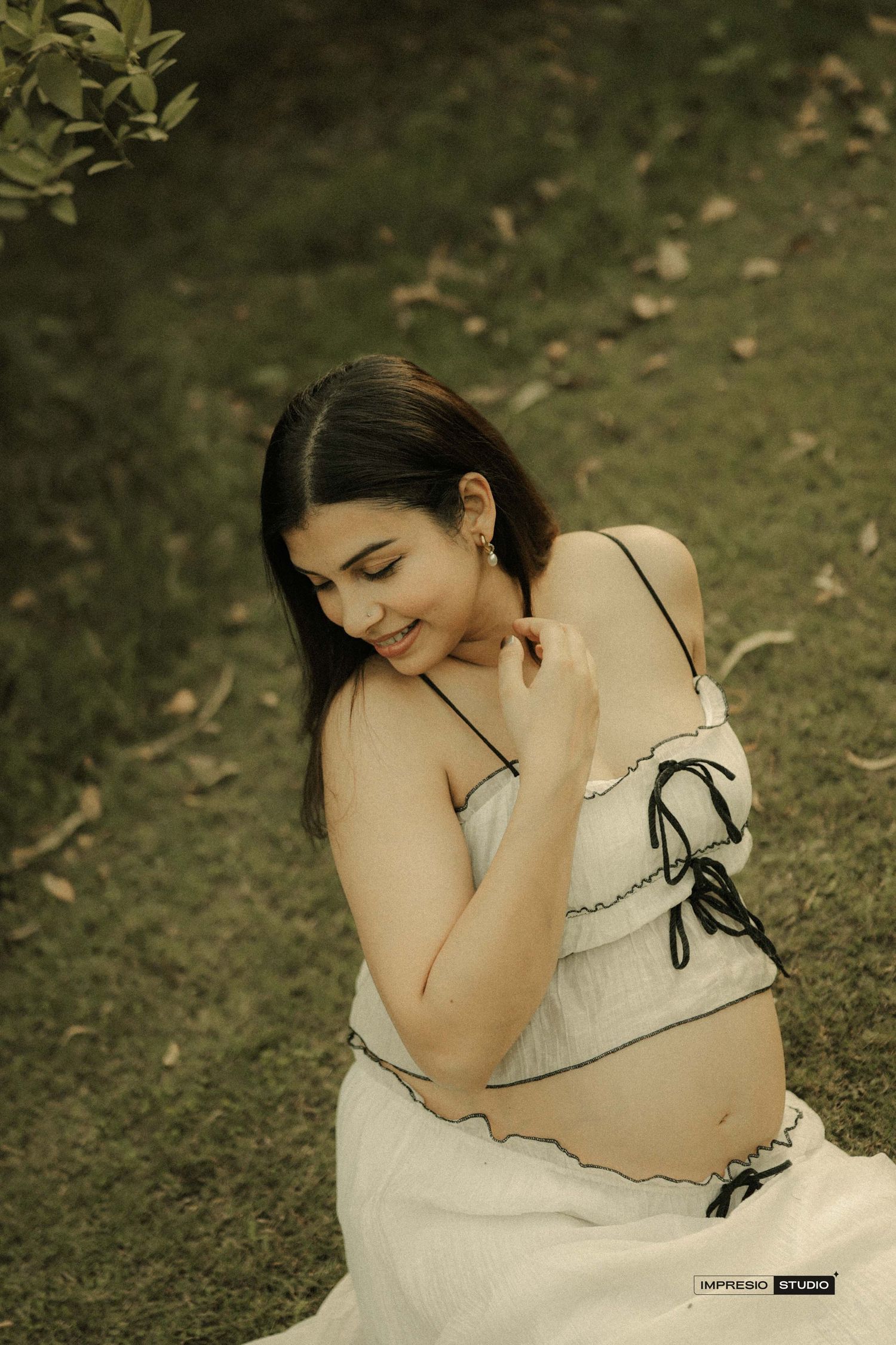 Pregnant woman in white crop top sits on grass, smiling serenely during an outdoor maternity photoshoot.