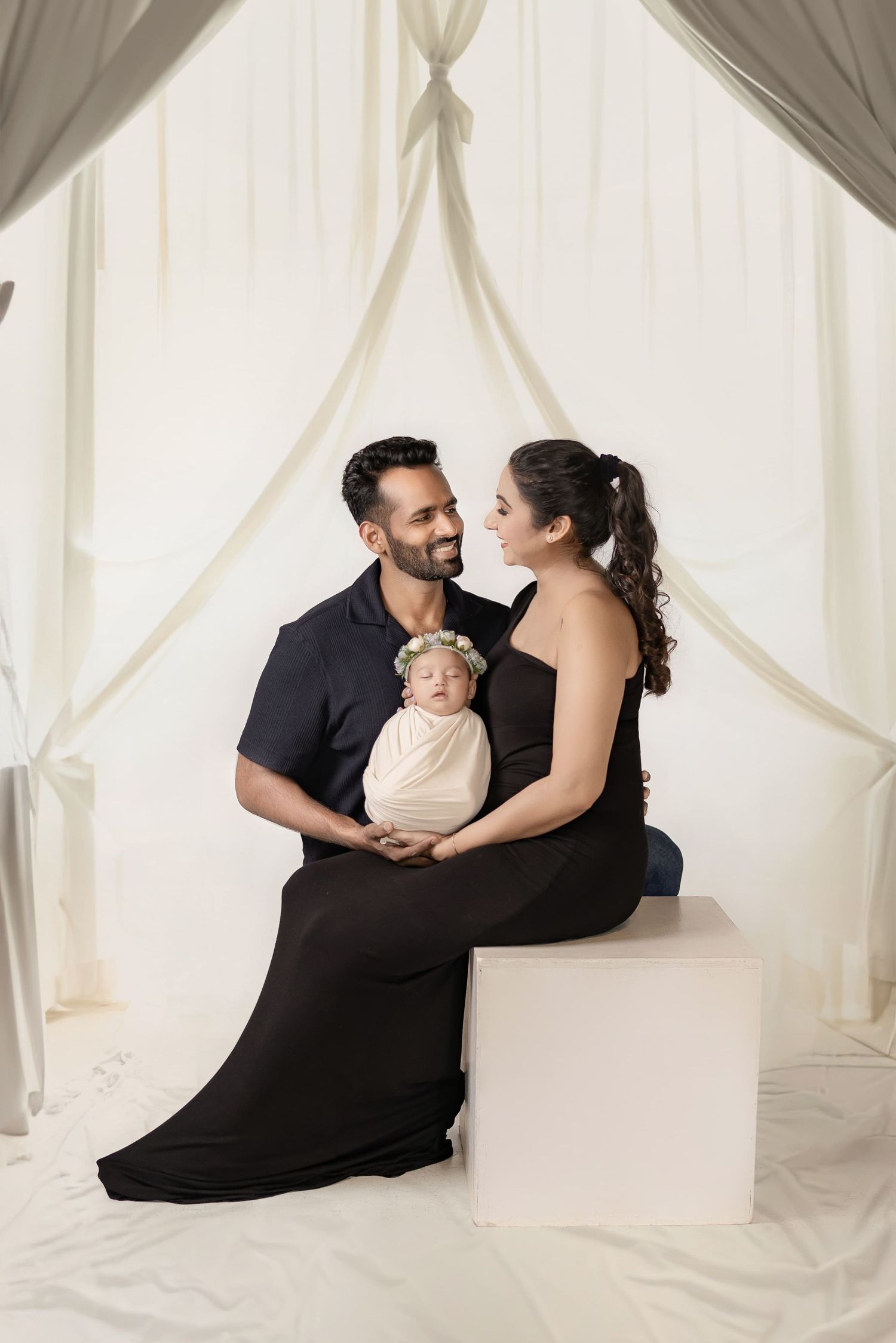 A couple in black outfits gazes lovingly at each other while holding their swaddled newborn baby in a white studio.