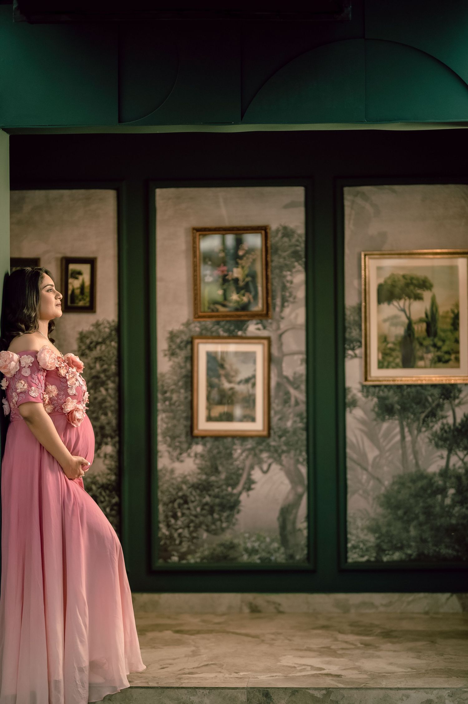 Pregnant woman in pink off-shoulder gown stands in art gallery with framed landscape paintings on decorative wall.