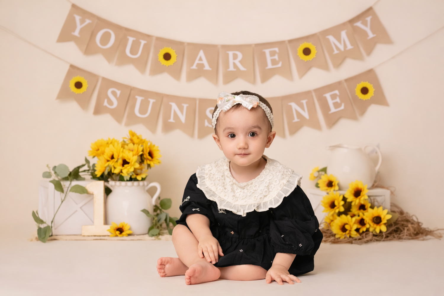 Baby in black dress sits on cream backdrop with 'You Are My Sunshine' banner and yellow sunflowers.