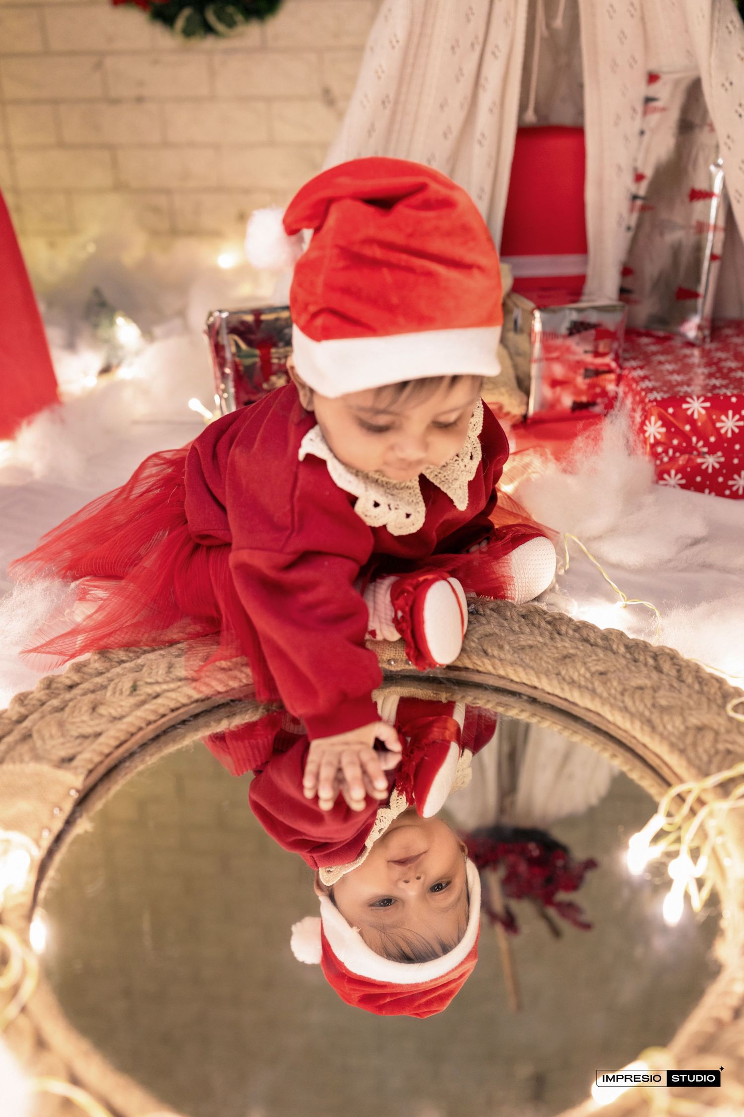 A baby in a red holiday outfit peers into a reflective ornament creating a mirrored holiday scene.
