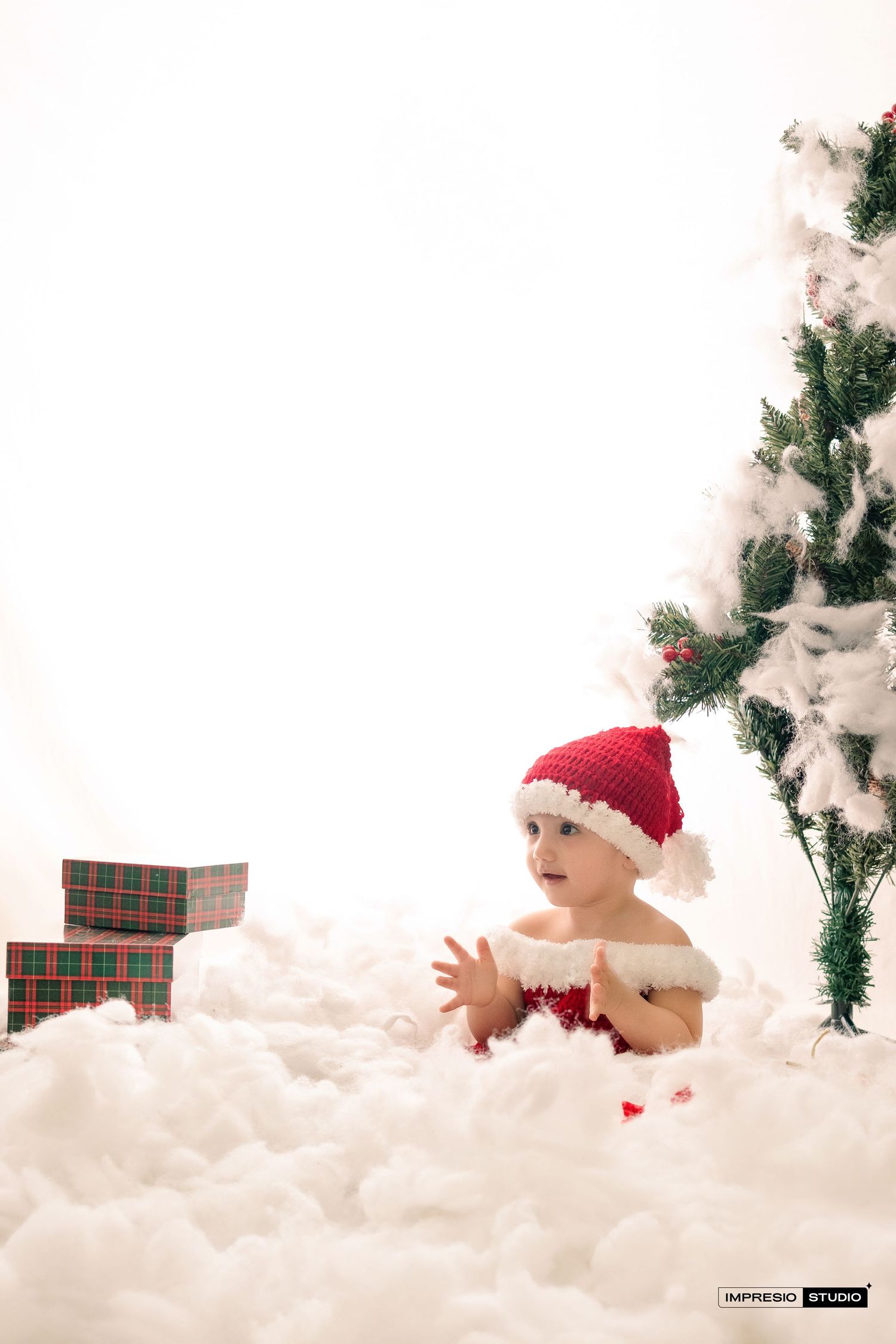 Baby wearing red Santa hat sits in white fluffy snow setting with Christmas tree and plaid presents in background.