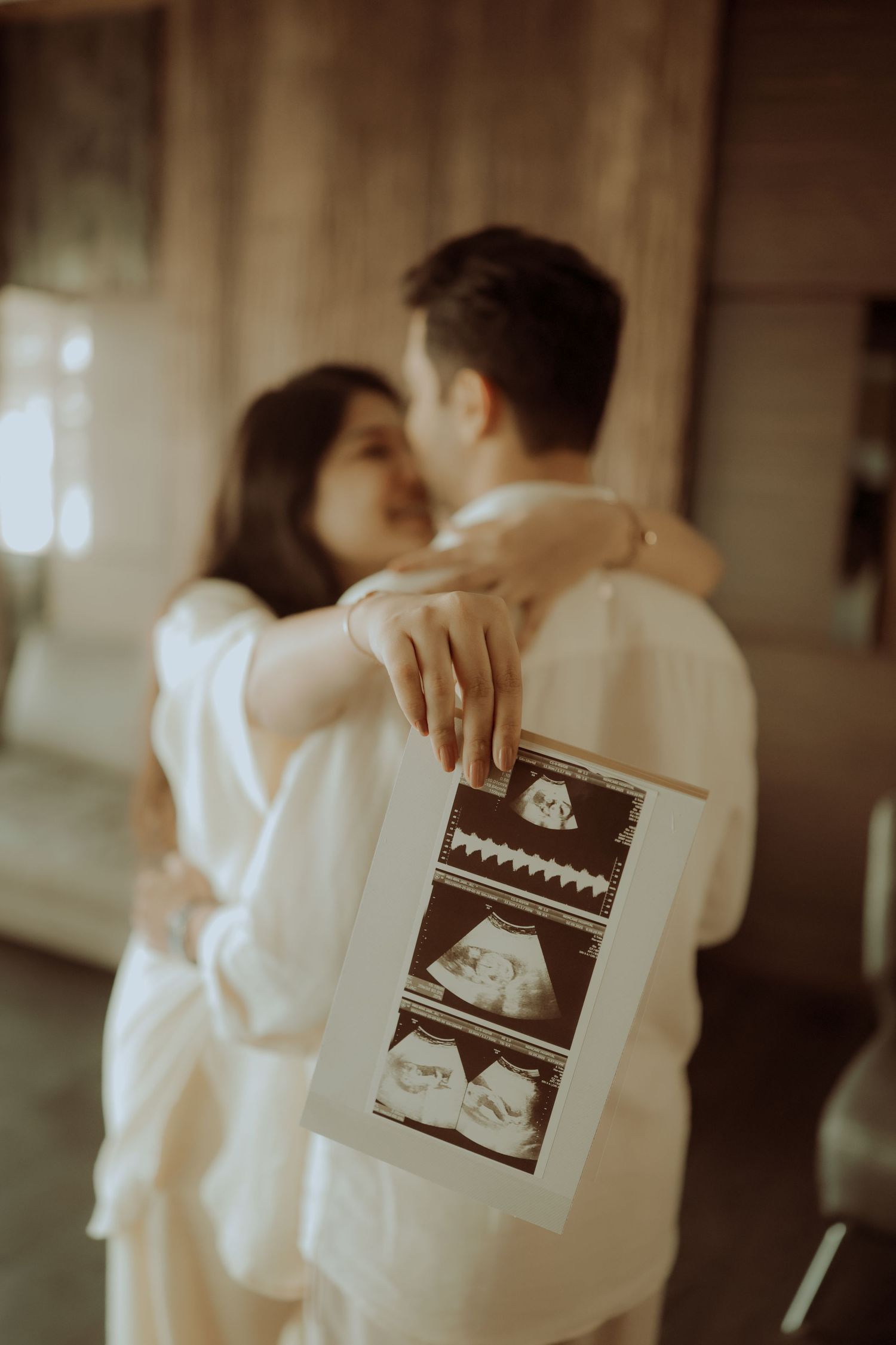 Couple embracing and kissing while holding ultrasound sonogram photos, announcing pregnancy in soft warm light.
