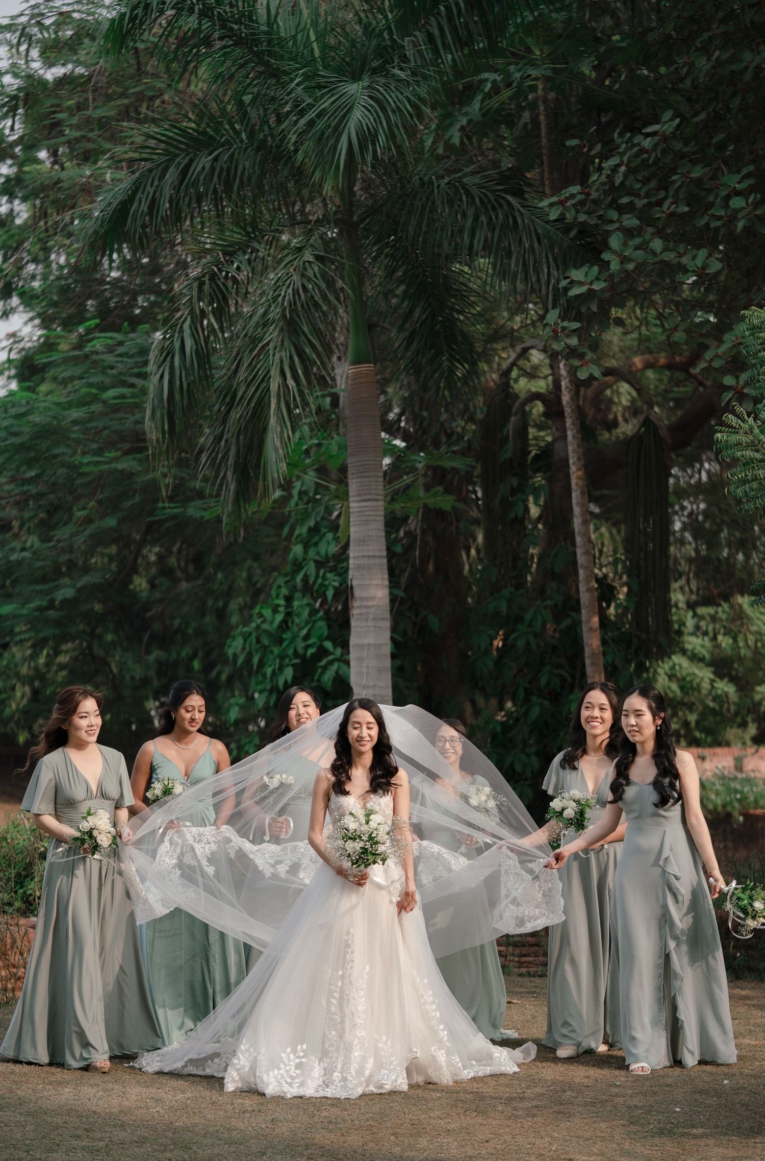 Bridal party in sage green bridesmaid dresses poses with bride in white gown holding bouquets in outdoor garden setting.