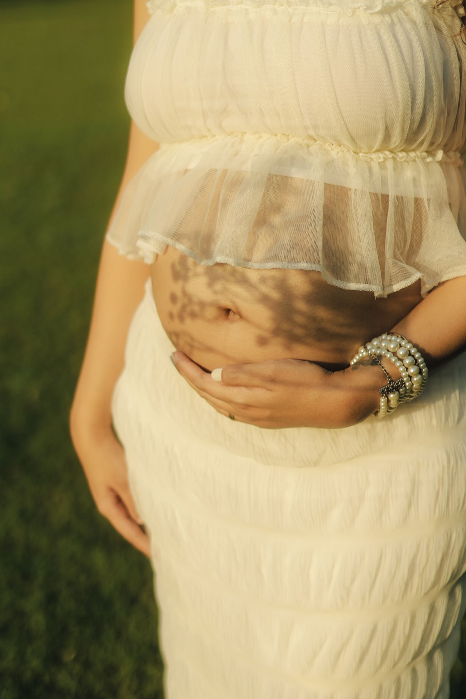 Pregnant woman in white dress cradling baby bump outdoors, wearing pearl bracelets, soft golden light on green grass.