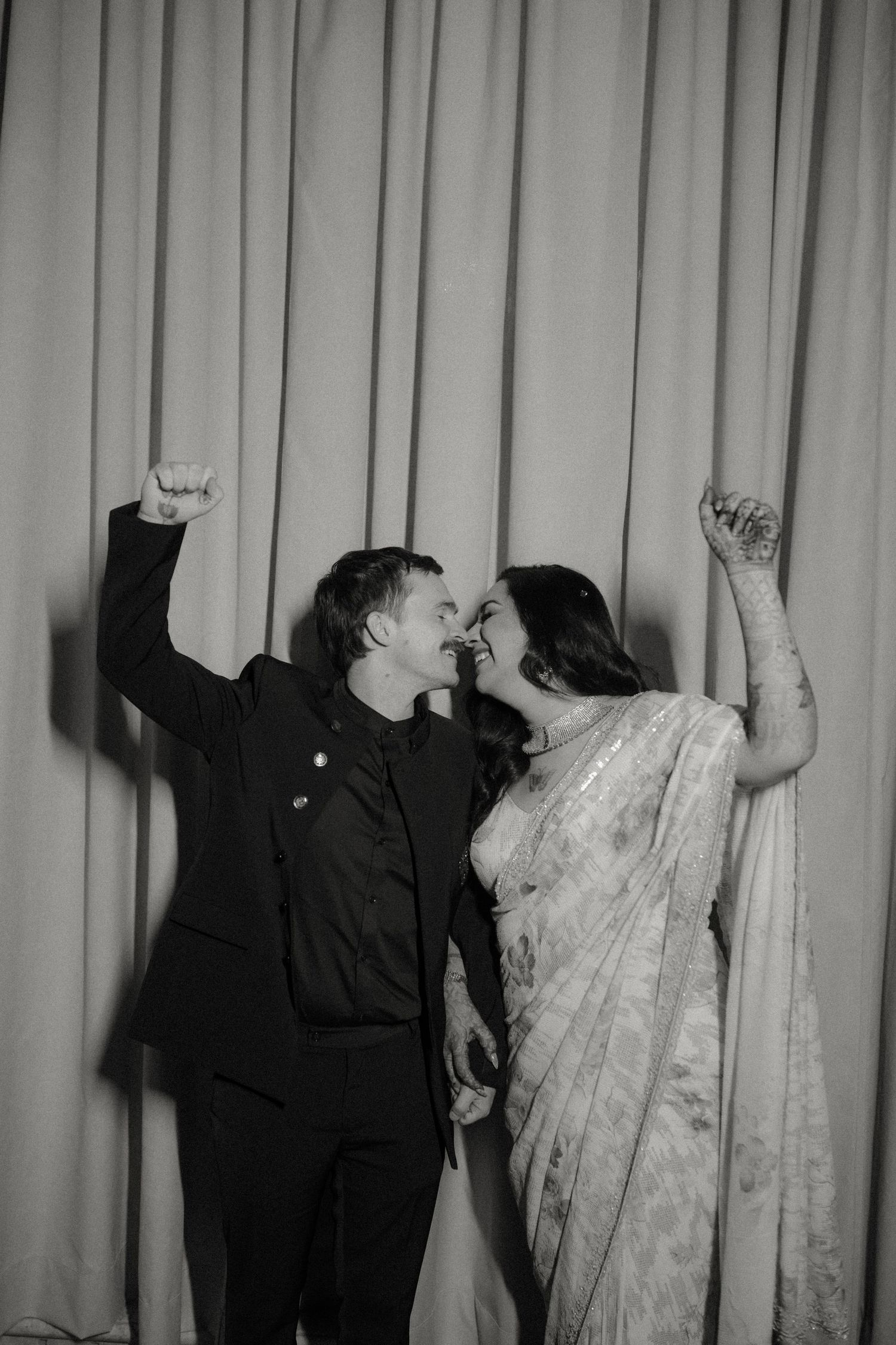 A joyful couple in western and traditional Indian attire dance together with raised arms against a curtained backdrop.