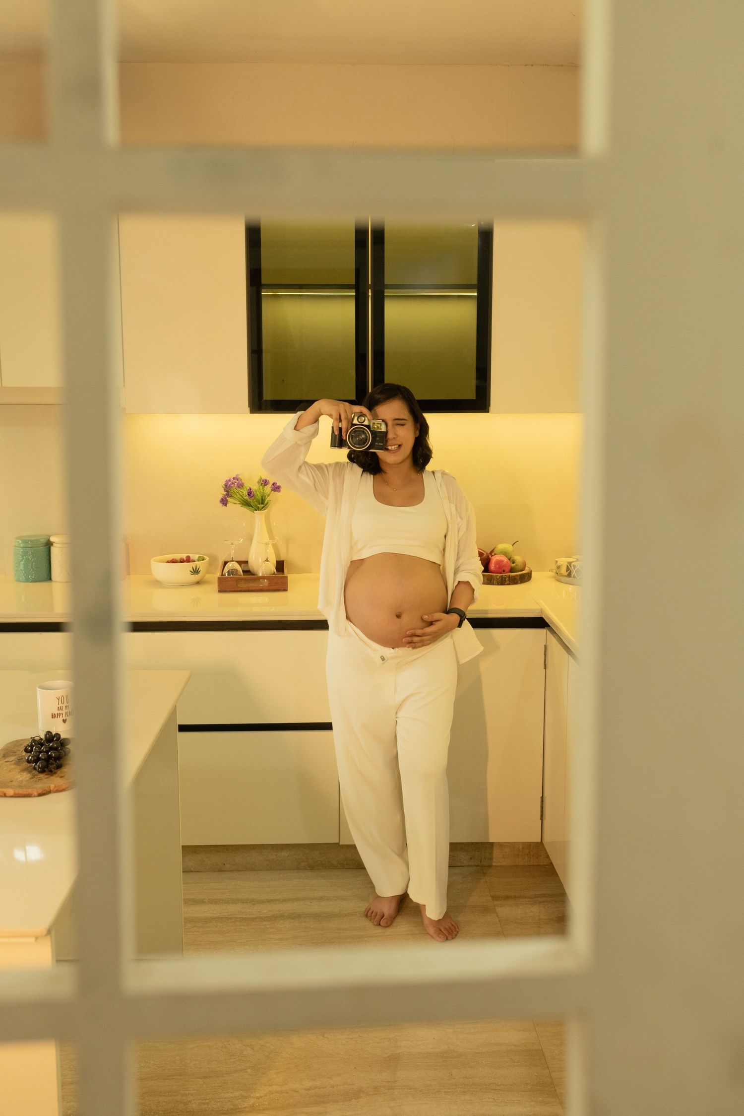 A maternity photoshoot in white loungewear standing in a modern kitchen with warm lighting viewed through a window pane.