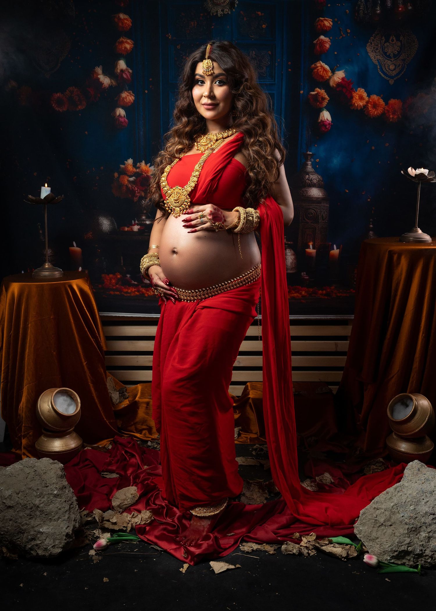 Pregnant person in traditional red Indian sari and gold jewellery poses against dark blue backdrop for maternity photography