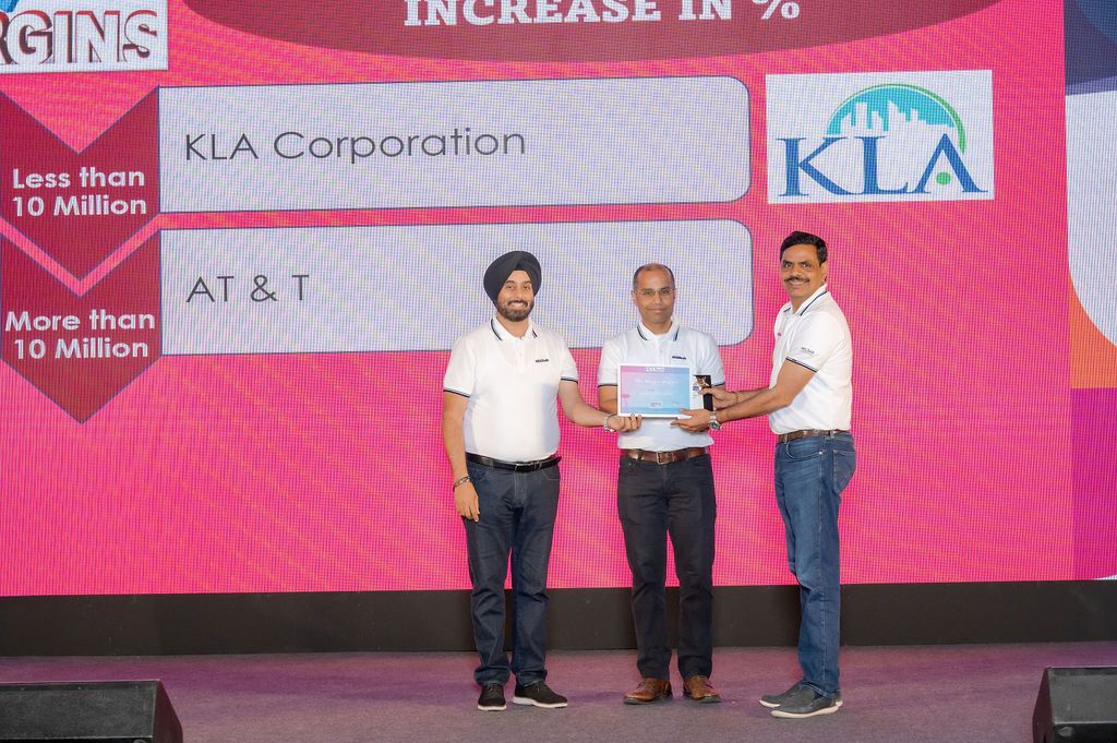 Three men in white shirts stand on stage presenting an award plaque at a corporate ceremony, with KLA Corporation and AT&T on the screen.