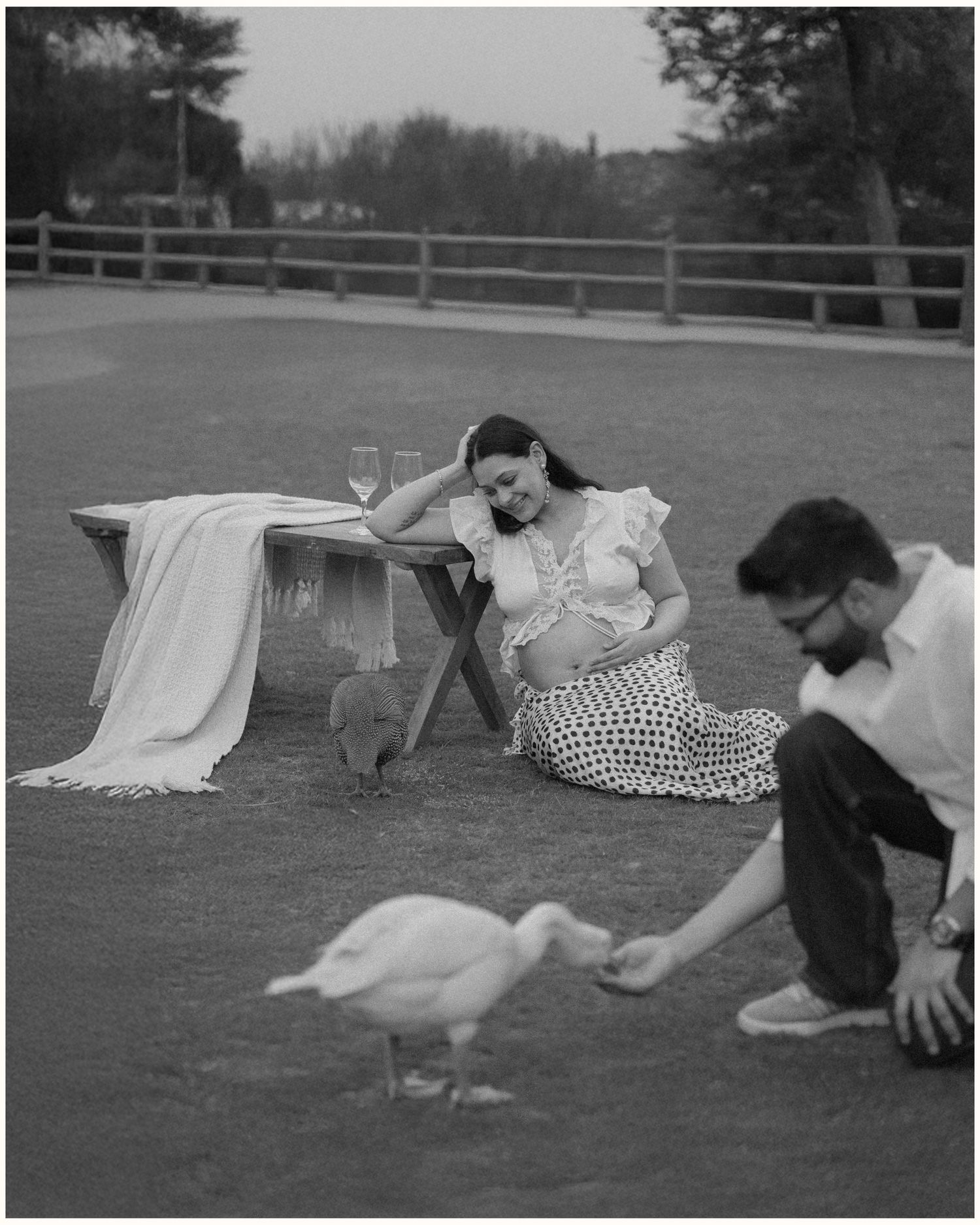 Black and white maternity photo of a pregnant woman sitting on grass while a man feeds a duck nearby.