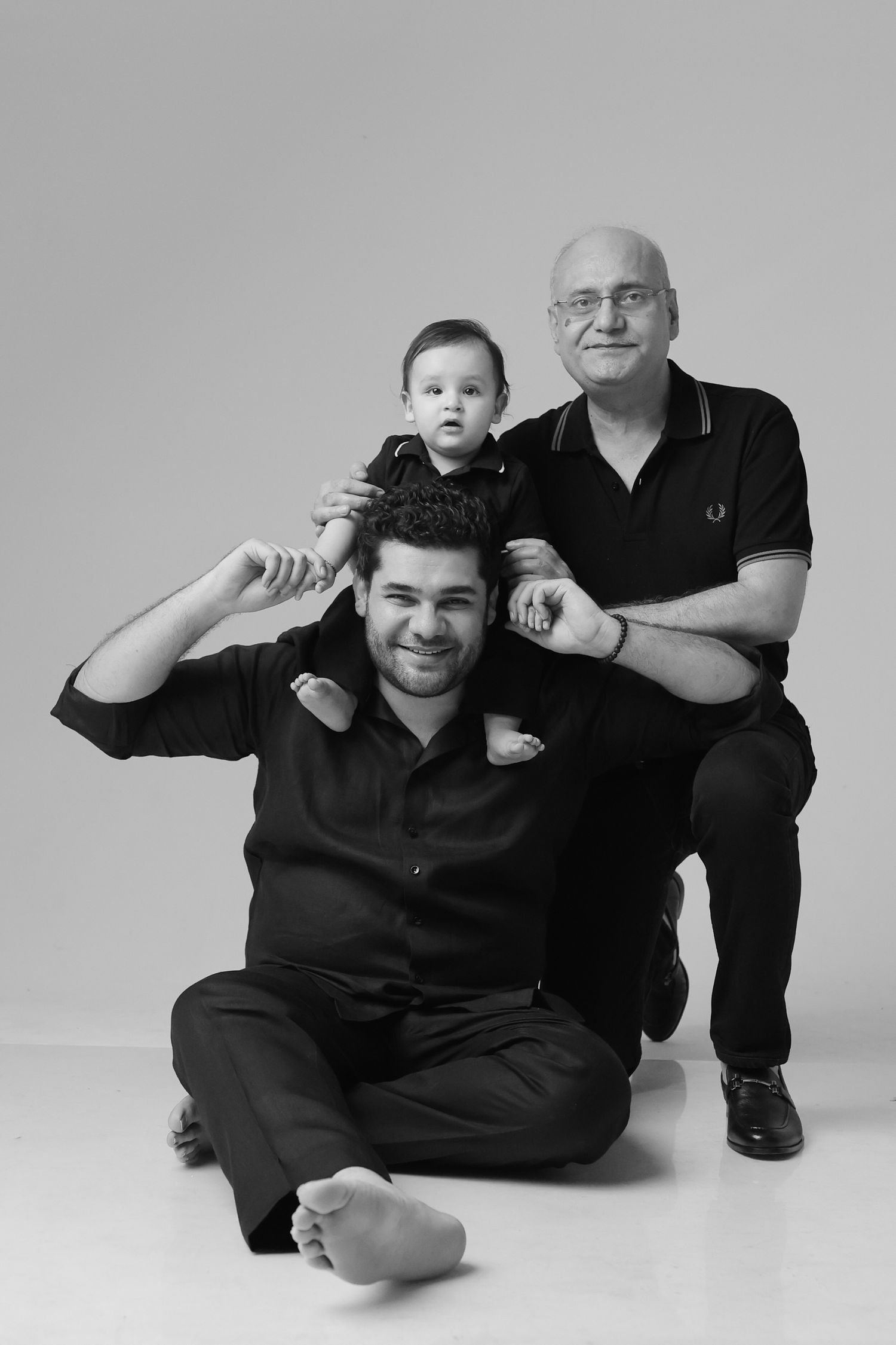 Black and white studio portrait of three generations, grandfather, father, and baby sitting together smiling.