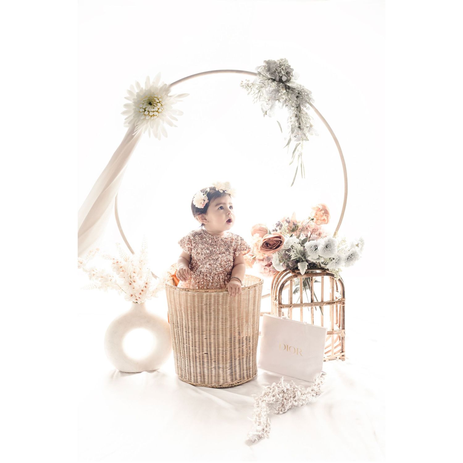 Baby girl in floral headband sits in wicker basket surrounded by dried flowers, birdcage, and circular floral hoop backdrop.