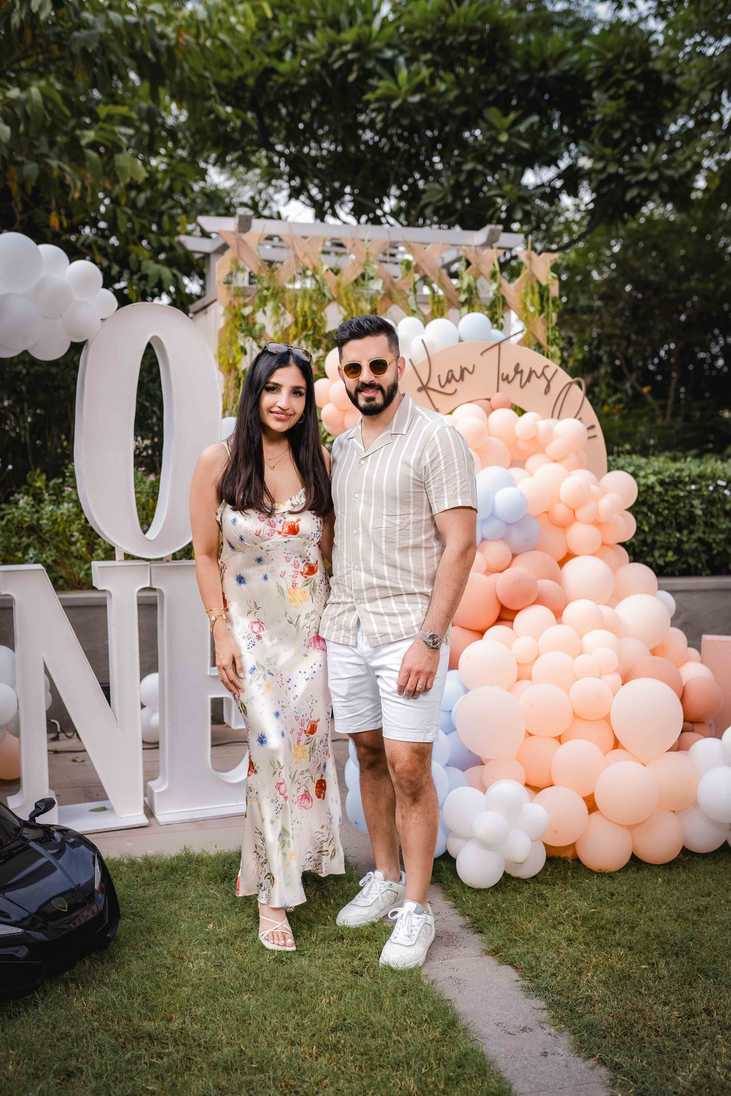 Couple posing at a first birthday party with balloon decorations and ONE sign outdoors.