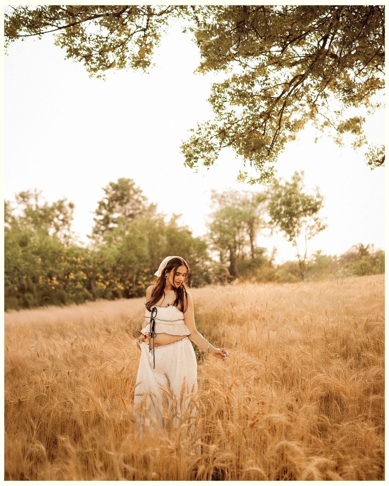 Woman in white outfit standing in golden wheat field surrounded by trees during golden hour.
