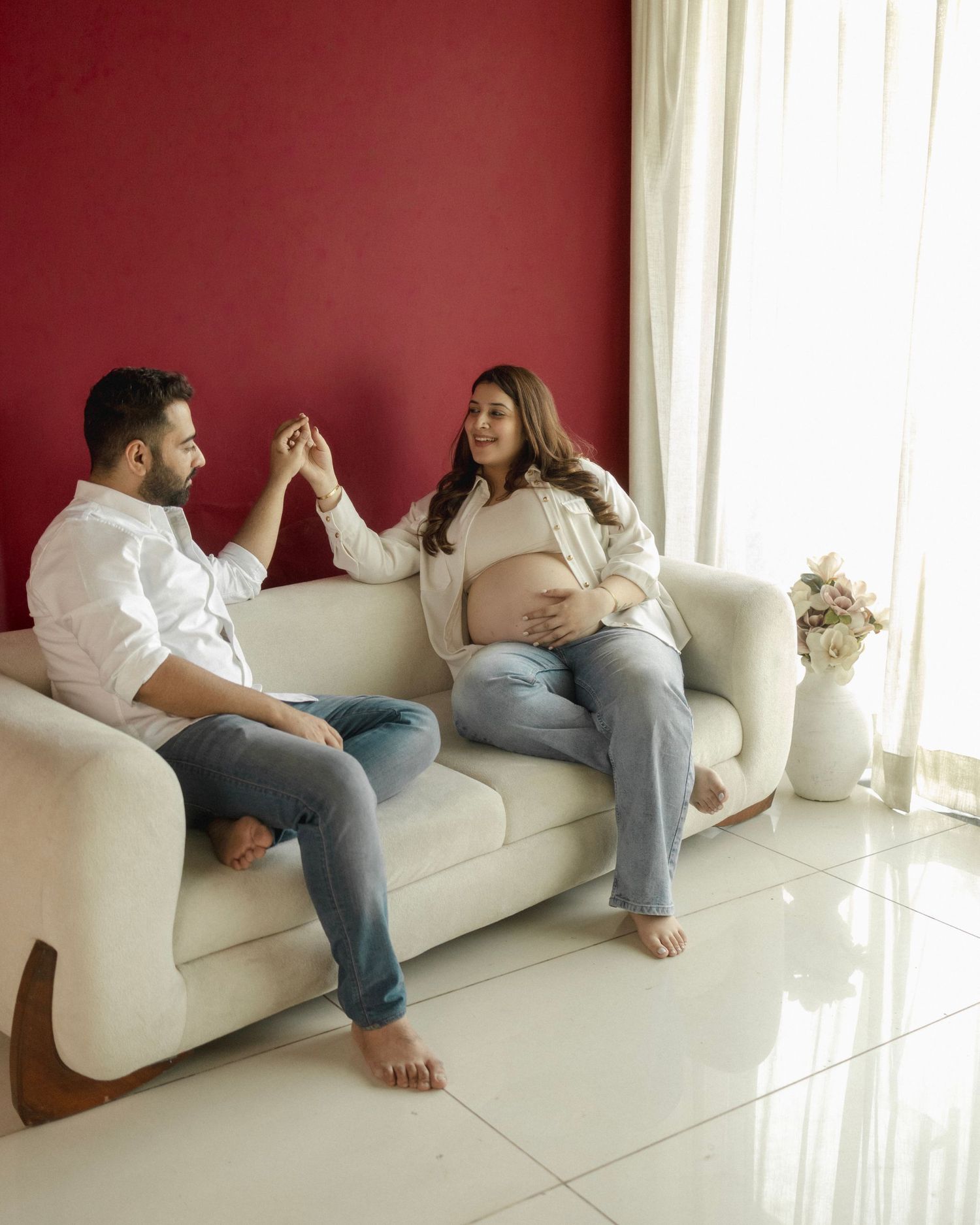 Pregnant woman and man sitting on white sofa, holding hands and smiling, with red wall background.