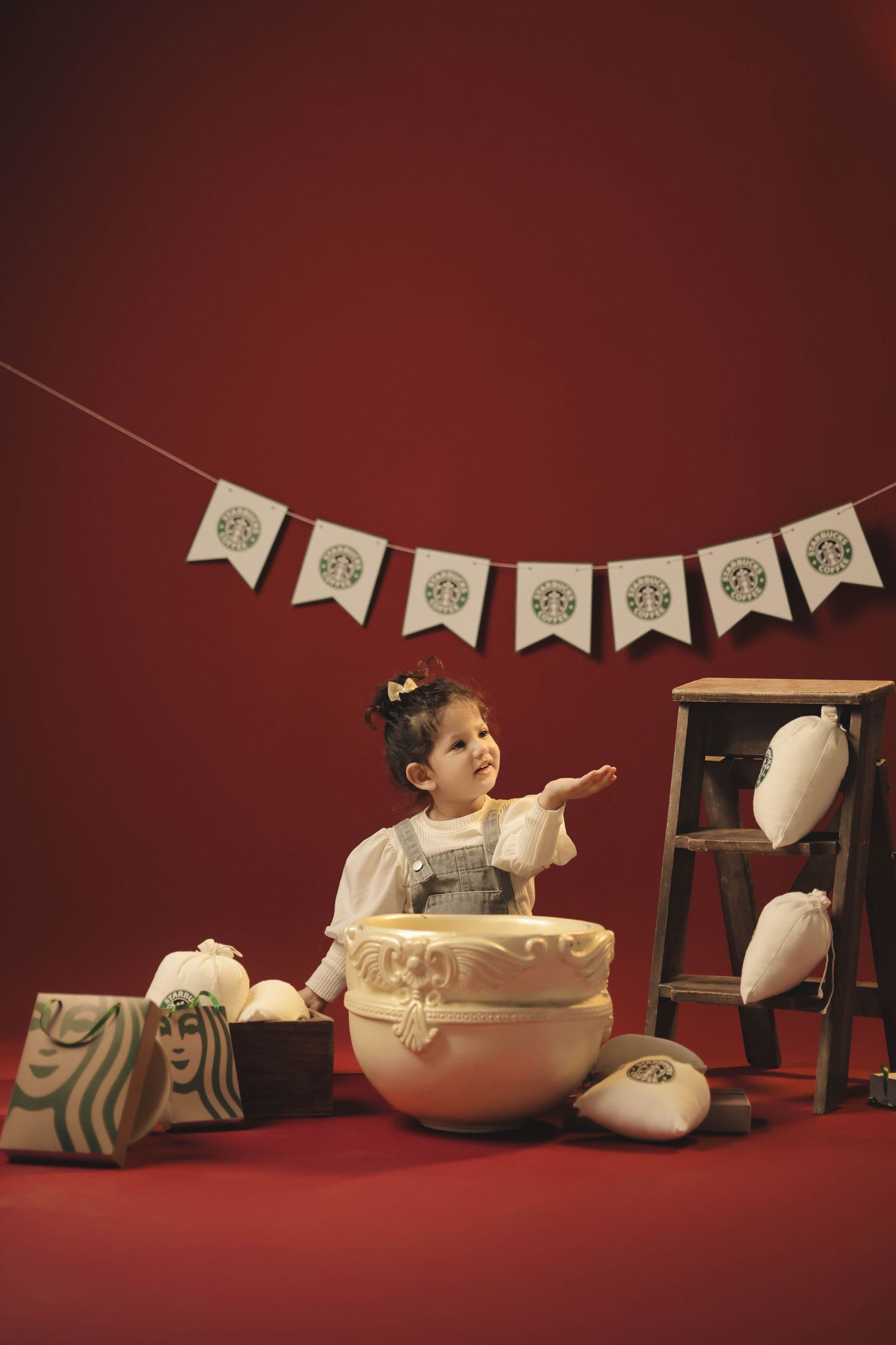 A young girl sits beside a large decorative drum with Starbucks-themed bunting and props on a red background.