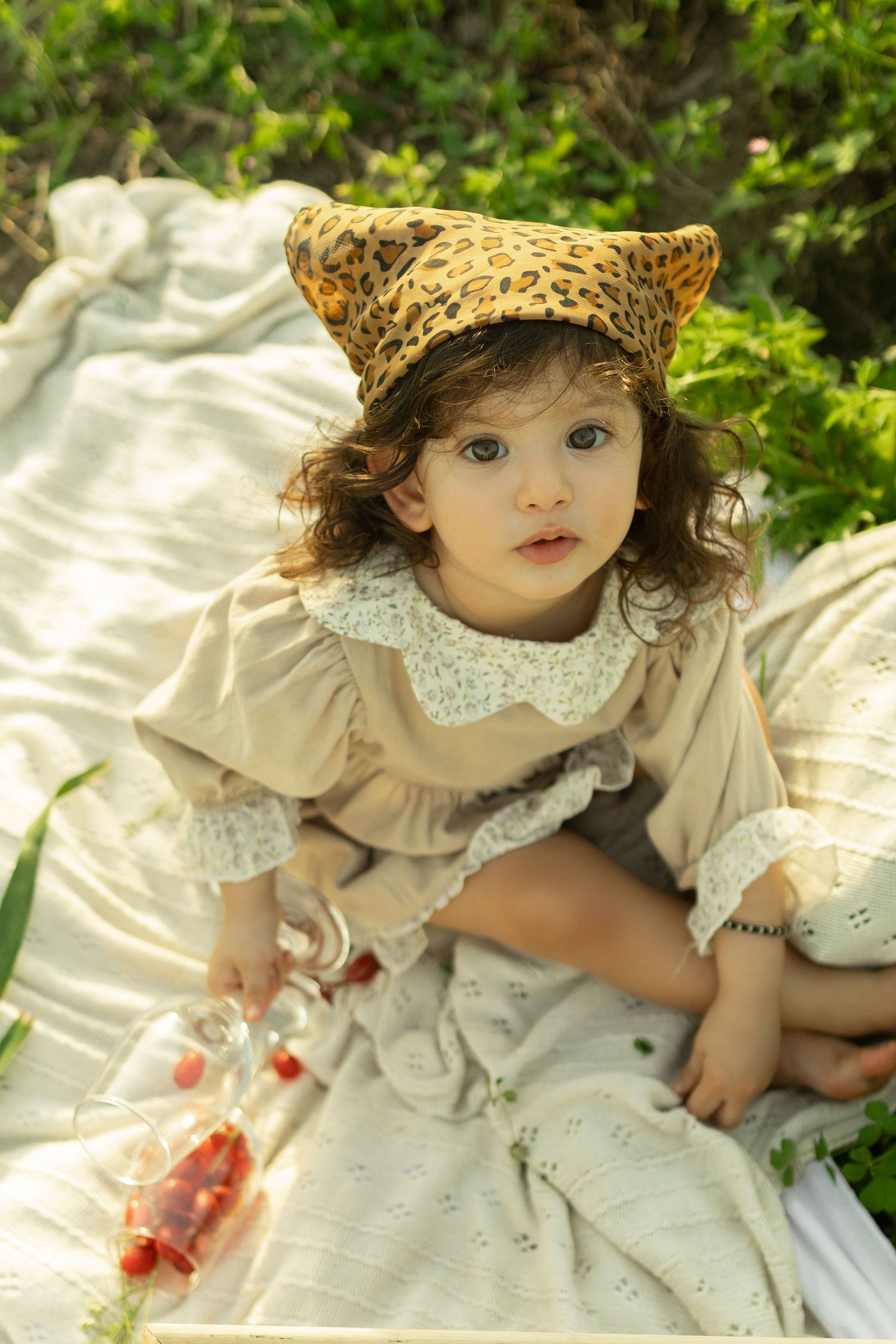 Toddler girl in beige lace dress and leopard print hat sitting on white blanket outdoors holding strawberries.