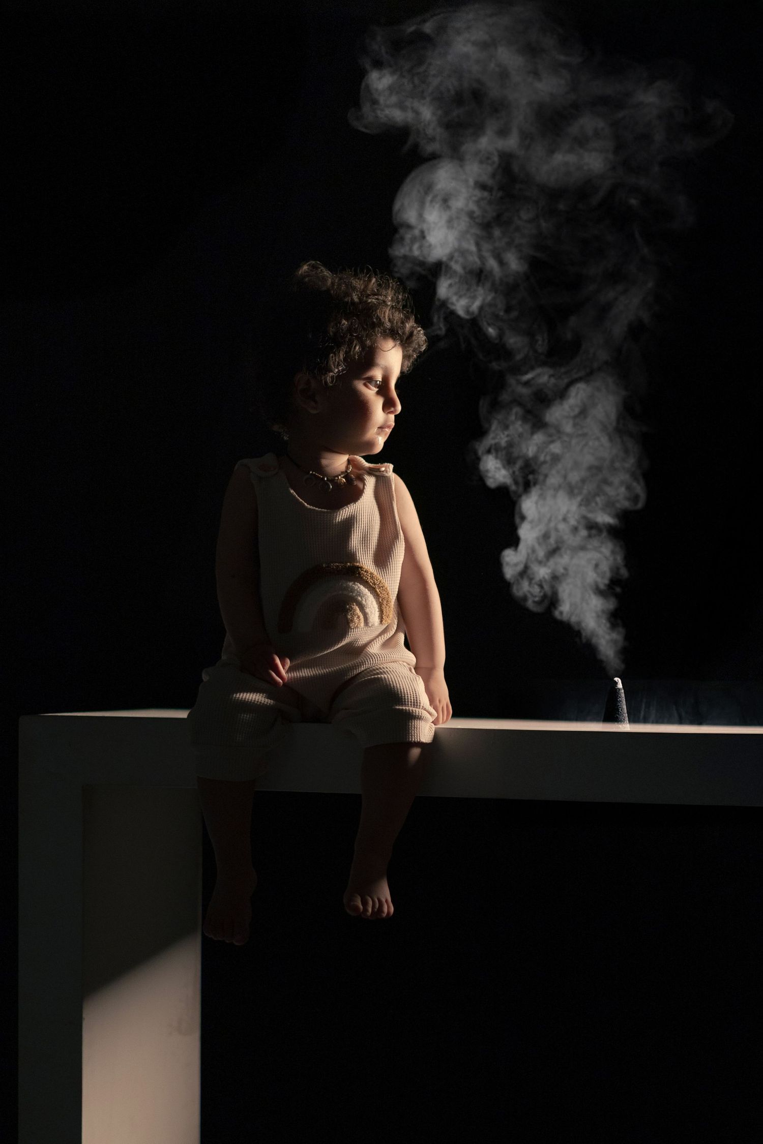 Young child sitting on white surface in dark studio with smoke rising beside them.