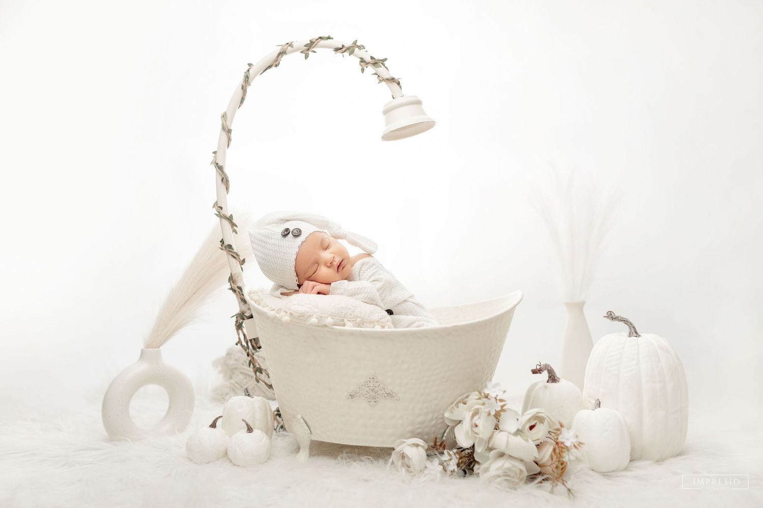 Newborn baby sleeping peacefully in a white boat-shaped bassinet surrounded by white pumpkins and flowers in a dreamy setup.
