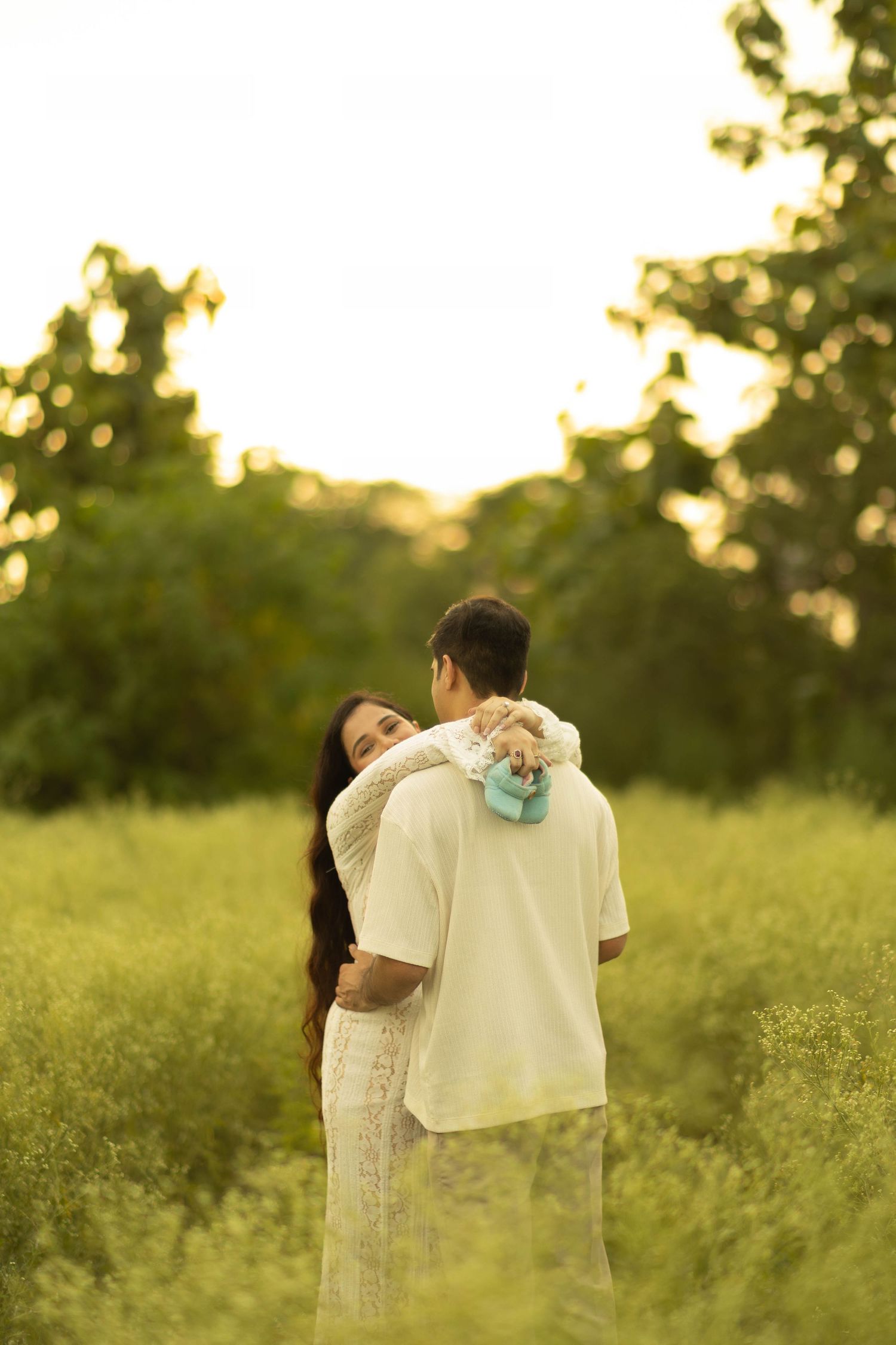 A couple embraces in a lush green field at sunset, with the man holding a baby wrapped in a blue blanket.