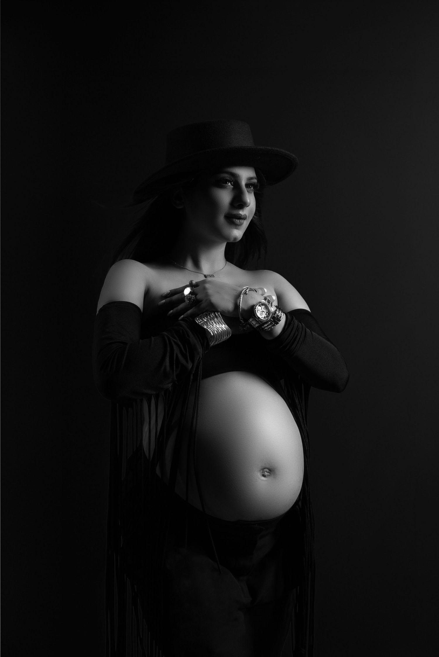 Black and white maternity photo of pregnant woman in wide-brim hat and off-shoulder top, cradling baby shoes on dark background.