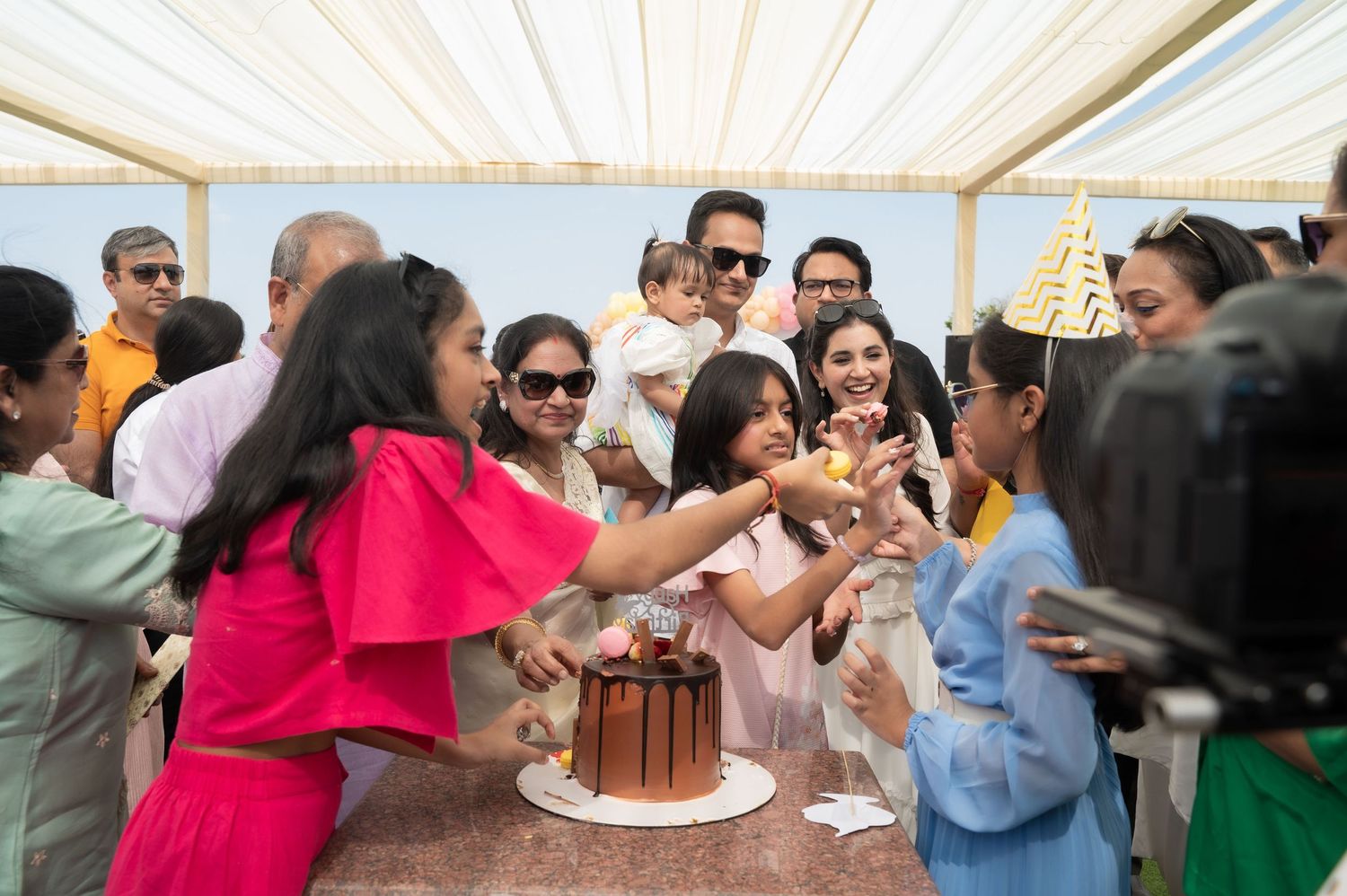 Group of people celebrating a birthday outdoors under a canopy, cutting a chocolate cake at a festive gathering.