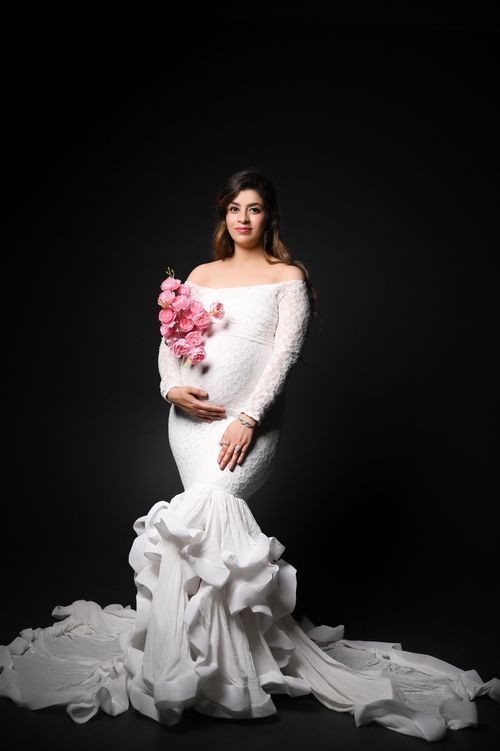 Dramatic maternity portrait in white mermaid gown with pink flowers against black backdrop.