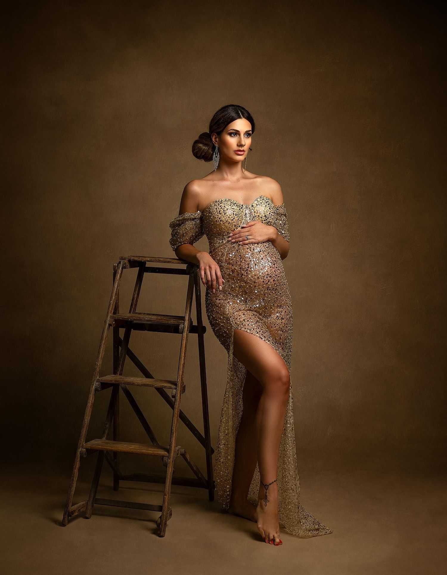 Artistic maternity photos with model posing elegantly on wooden chair in sequined dress.