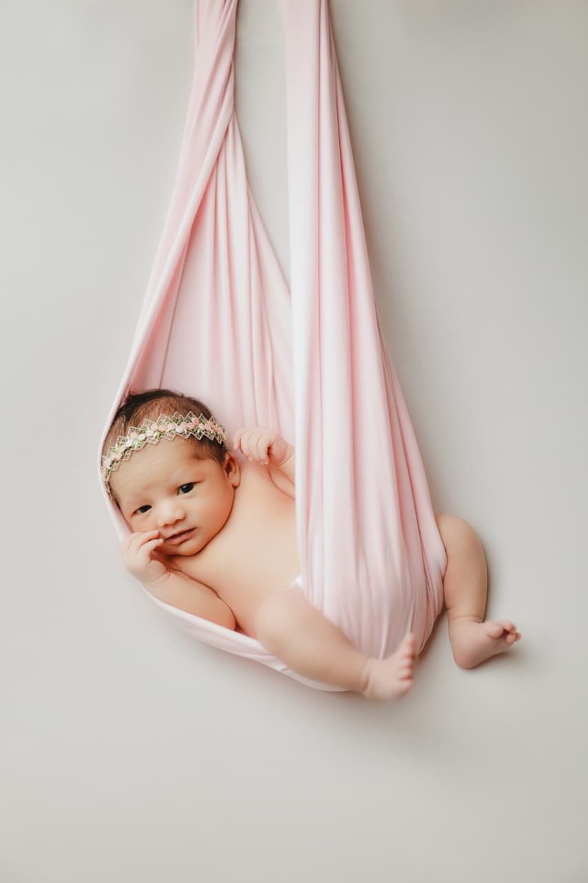 Peaceful newborn baby girl in a pink fabric hammock wearing a delicate gold headband.