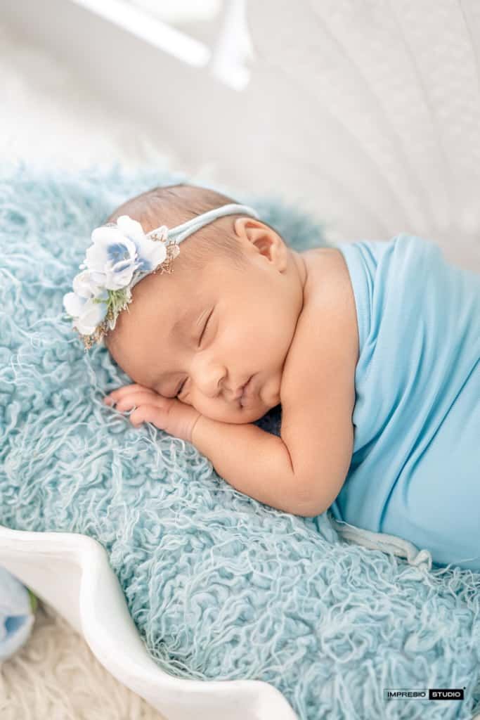 Sleeping newborn baby girl wrapped in light blue swaddle with floral headband on soft teal fur blanket.