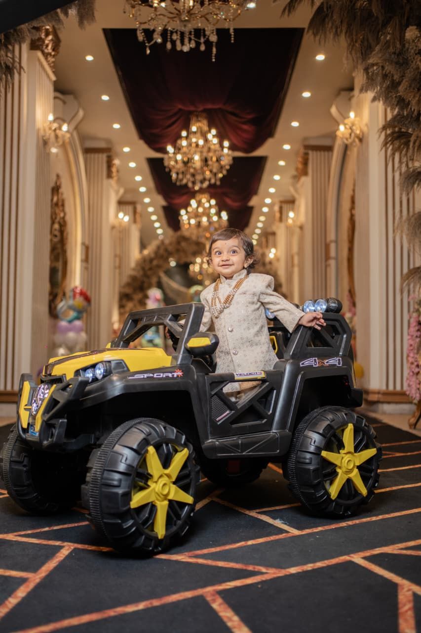 Young child smiling in a black and yellow ride-on toy SUV jeep inside an elegant decorated hall.