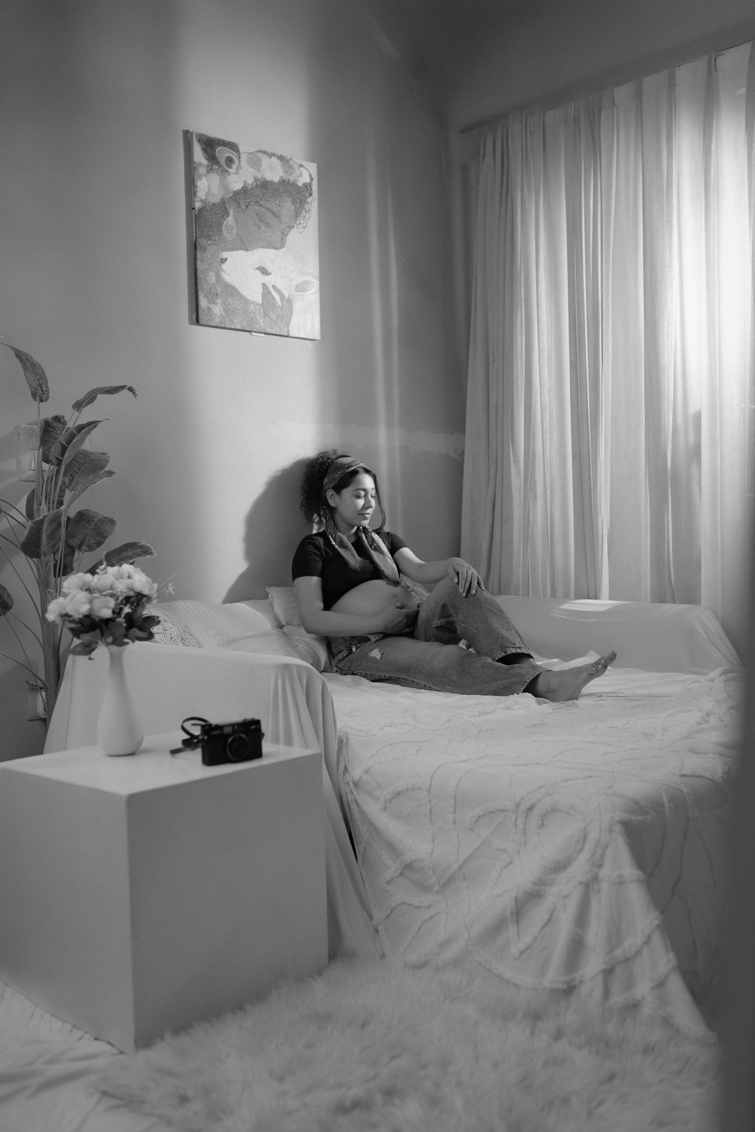 Black and white photo of a pregnant woman sitting on a bed in a cozy bedroom with curtains and plants.